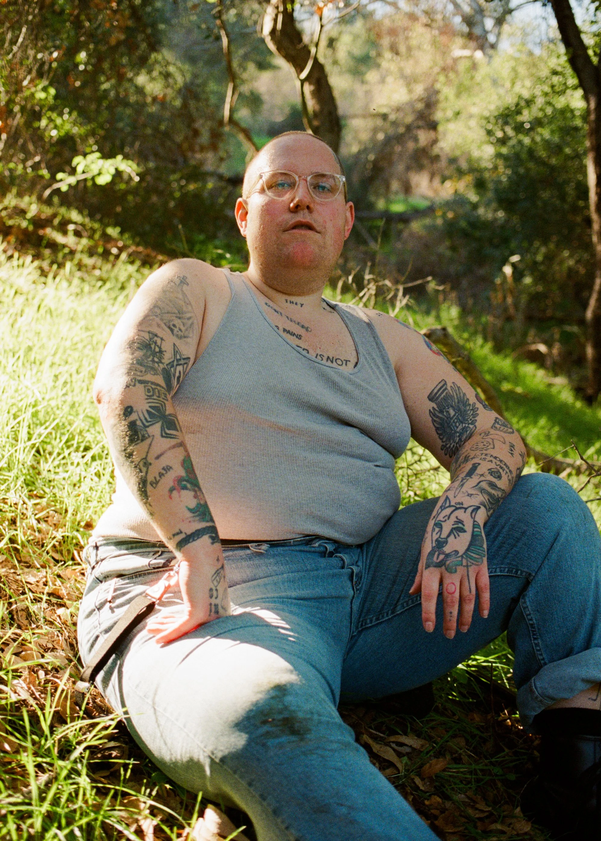 A nonbinary person with tattoos, wearing glasses, a sleeveless shirt, and jeans, sitting outdoors on grass and fallen leaves, with trees in the background. Shot by photographer Holly Stevens.