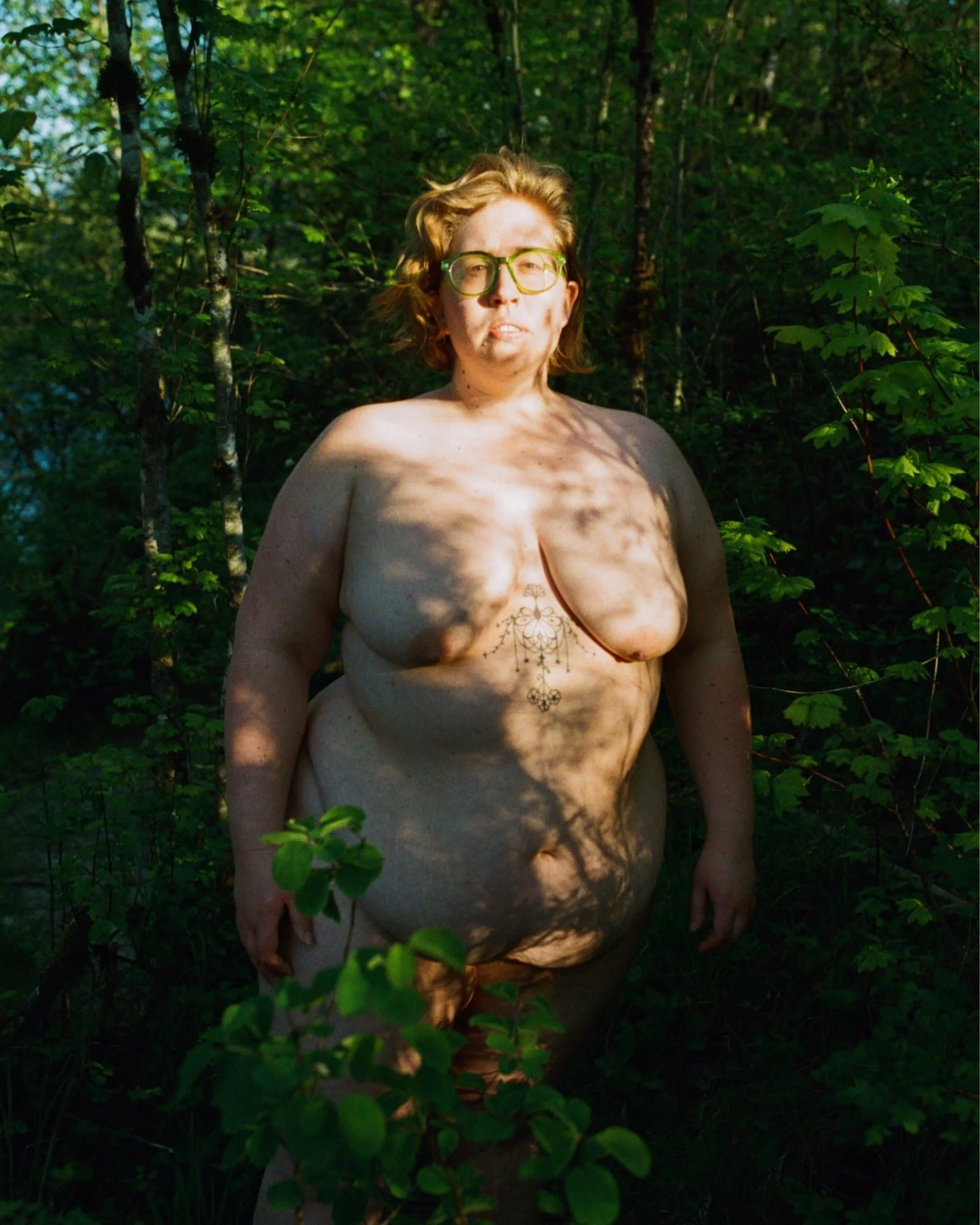 A nude woman with glasses and short blonde hair standing in a forest, with sunlight and shadows on her body through the leaves.