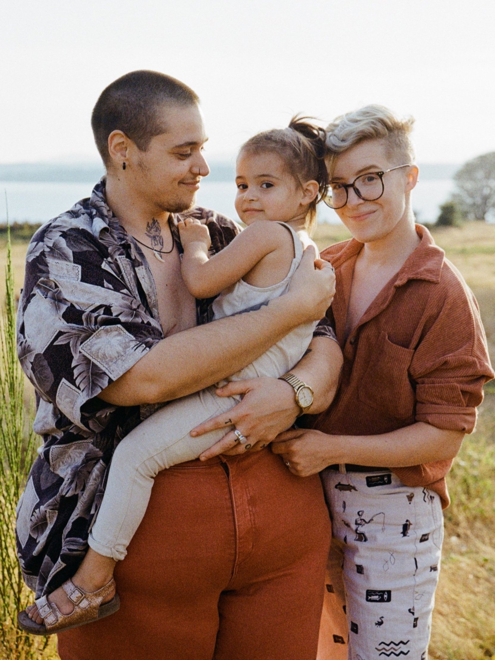 Two nonbinary parents stand together holding a young child who is smiling at the camera. Shot by portrait photographer Holly Stevens.