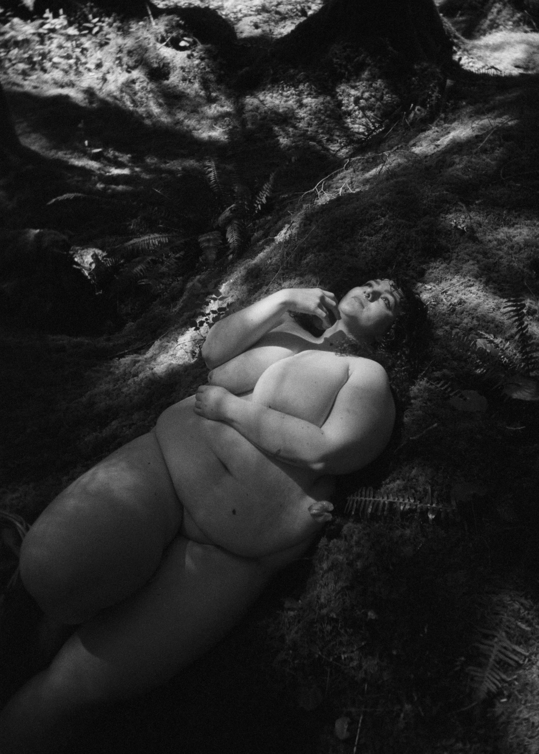 A black-and-white photo of a nude woman lying on a forest floor surrounded by moss and ferns, with her head turned upward and one hand touching her neck.