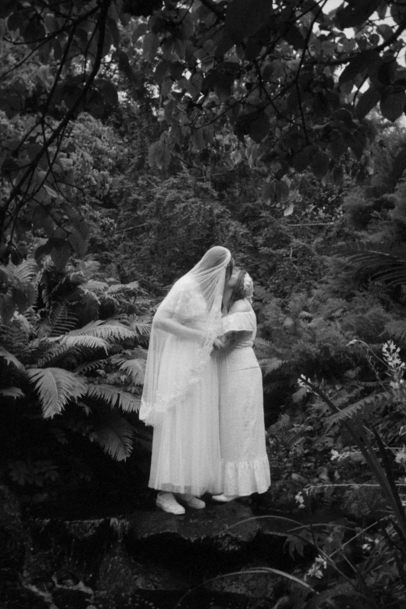 Two brides dressed in white stand together as they kiss in a lush forest. Shot by queer wedding photographer Holly Stevens.