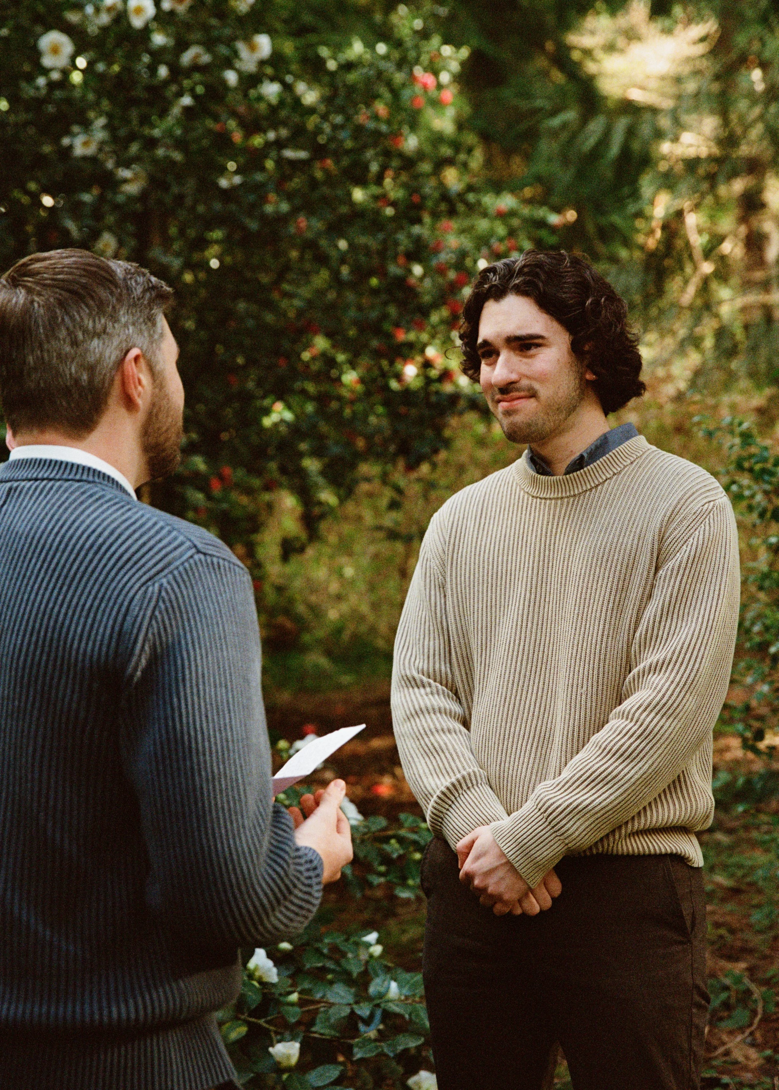 A man with curly dark hair and light skin is getting married to another man in a suit, outdoors among greenery and flowers.