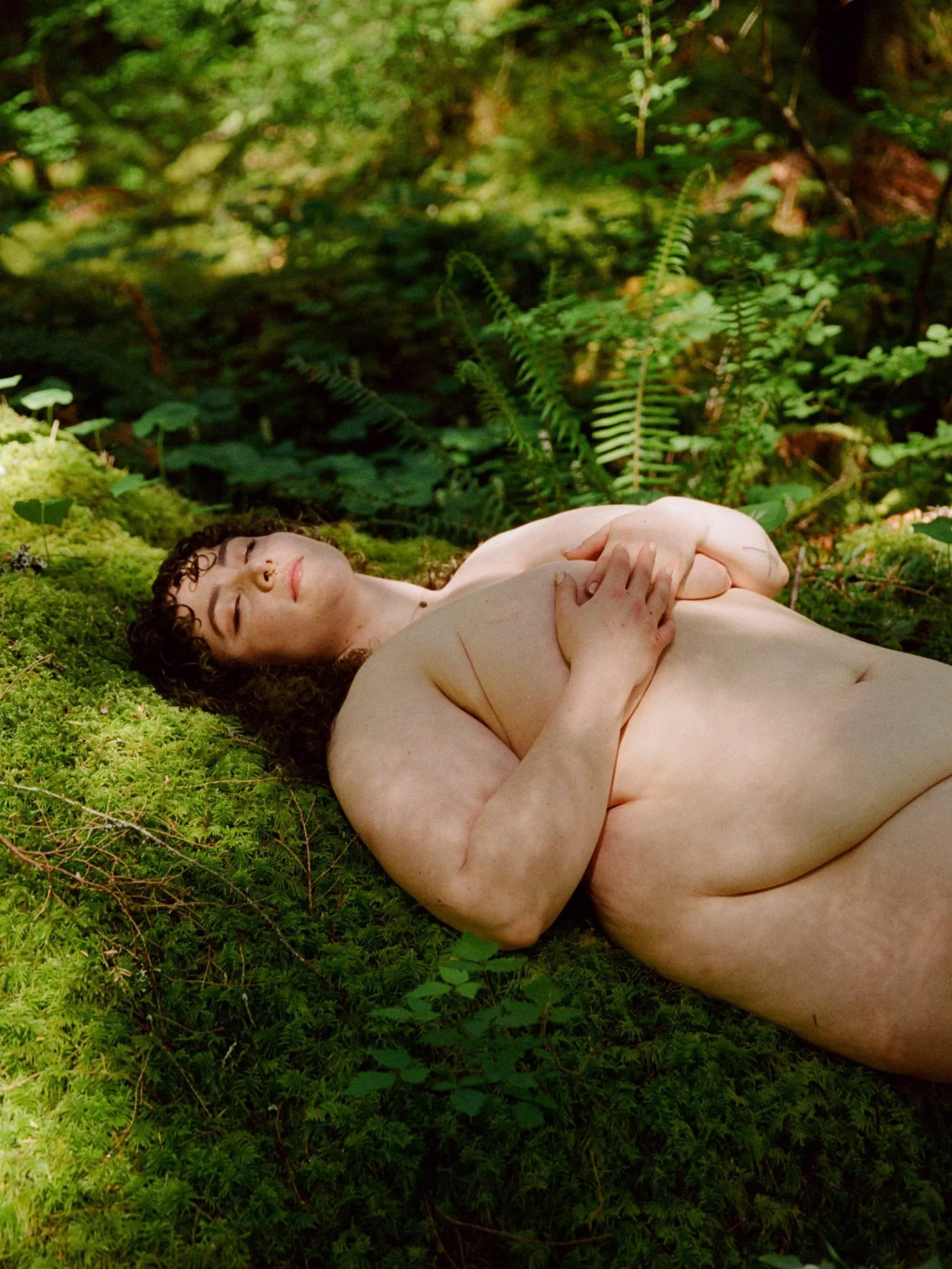 A nude woman in Seattle with curly dark hair and fair skin lying on green moss in a forest, with her eyes closed and arms crossed over her chest.
