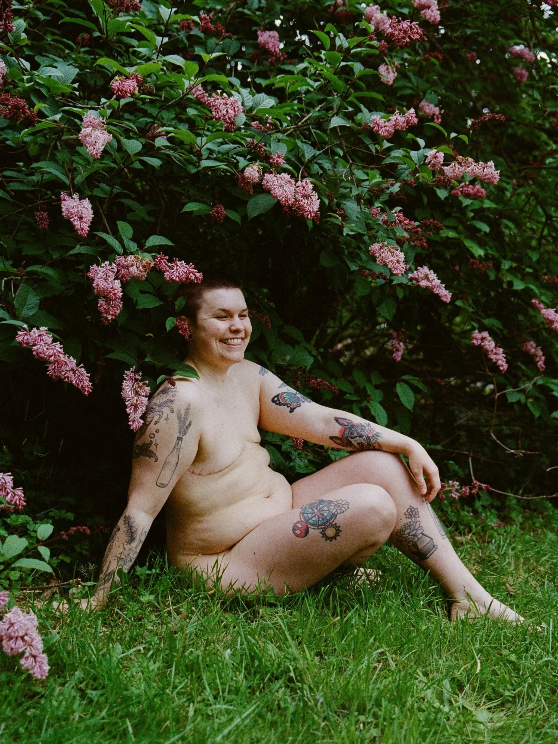 A nude woman with tattoos sitting on the grass in front of pink flowering bushes, smiling. Shot by queer photographer Holly Stevens.