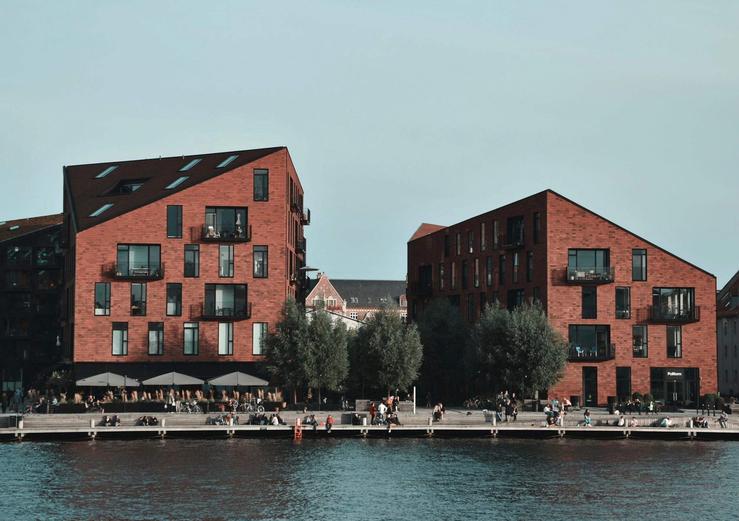 Moderne røde boligbyggerier ved en flod med gående og cyklende mennesker foran.