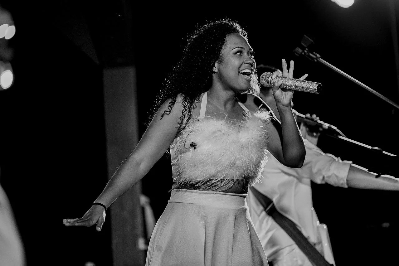 A woman singing into a microphone on a stage, smiling, with her arms outstretched, wearing a feathered top and a high-waisted skirt, with a dark background and stage lights.