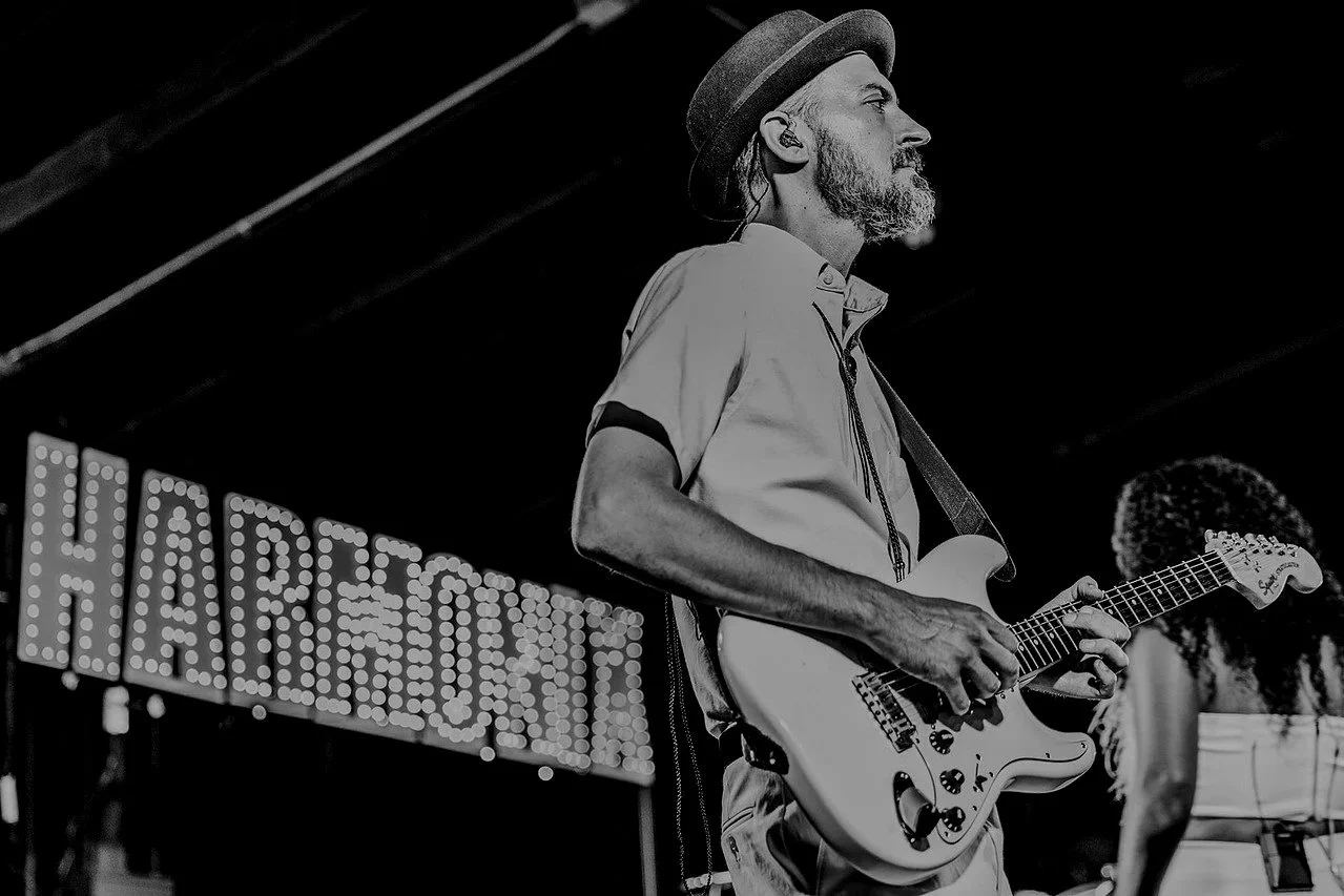 A man with a beard wearing a hat playing an electric guitar on stage, with a sign reading 'HAREWOOD' in the background.