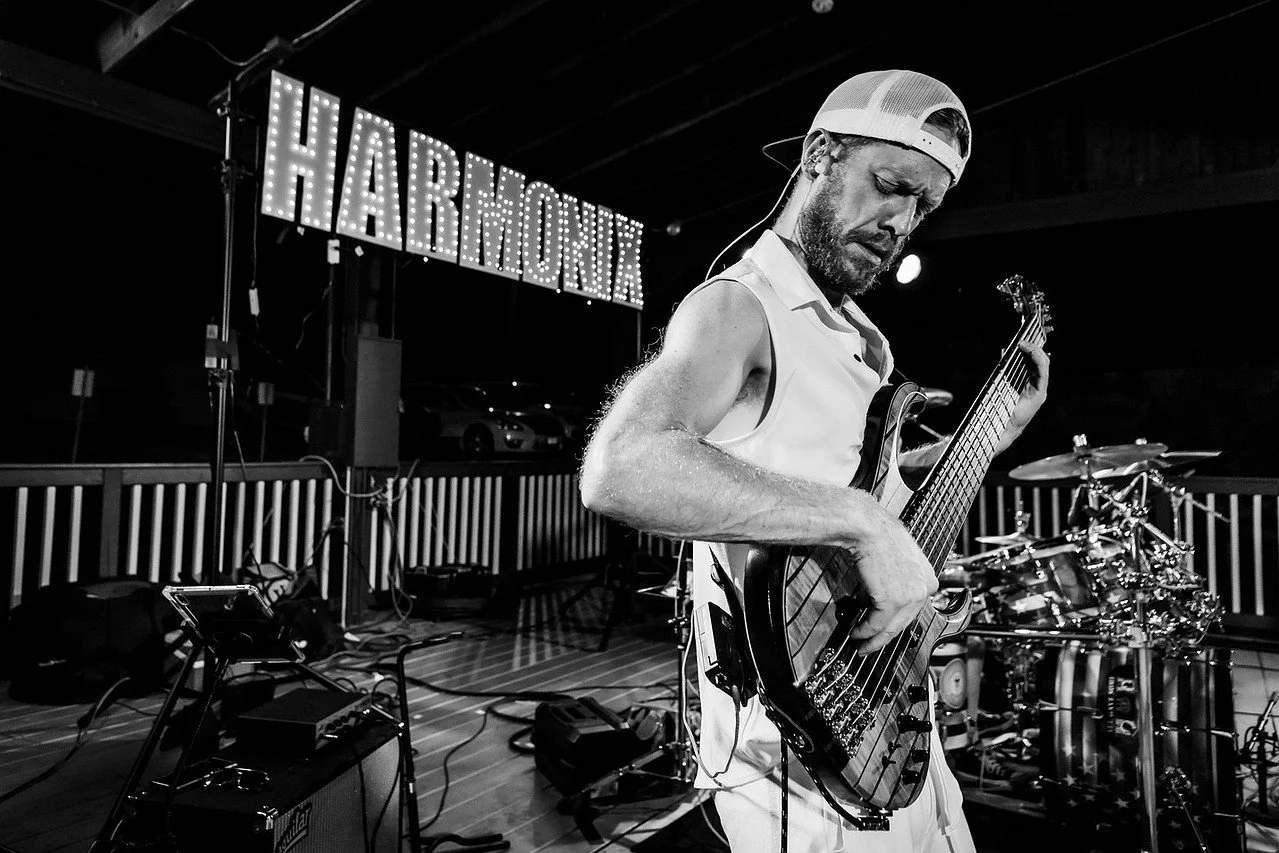 A man wearing a sleeveless shirt and baseball cap playing a bass guitar on a stage with drum equipment in the background, and a lit sign that says 'HARMONY' behind him.
