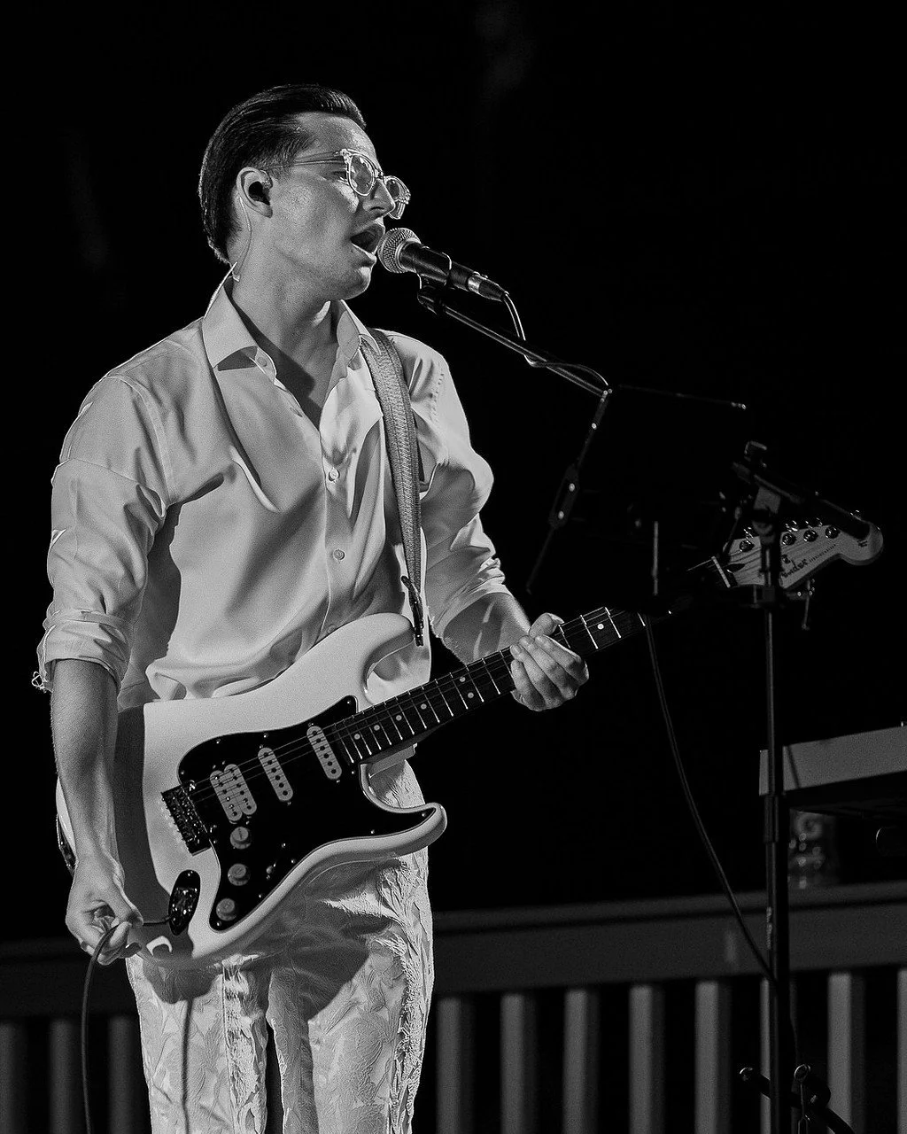 A black and white photo of a person singing into a microphone while playing an electric guitar on stage.