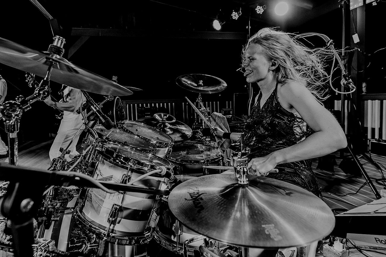 A woman playing a drum set passionately on stage, with hair flying and surrounded by musical equipment.
