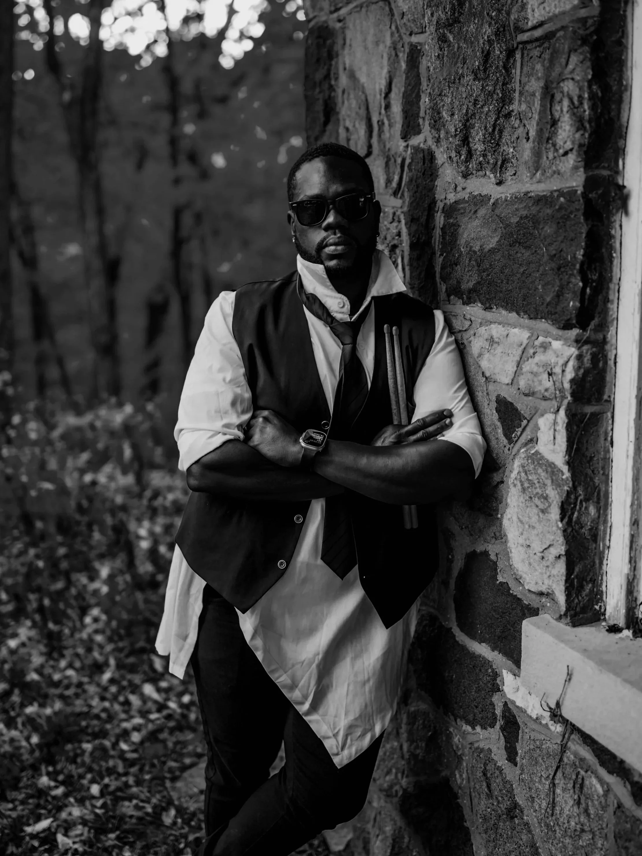 A man standing outdoors against a stone wall, wearing sunglasses, a vest over a shirt with rolled-up sleeves, and a tie, with crossed arms.