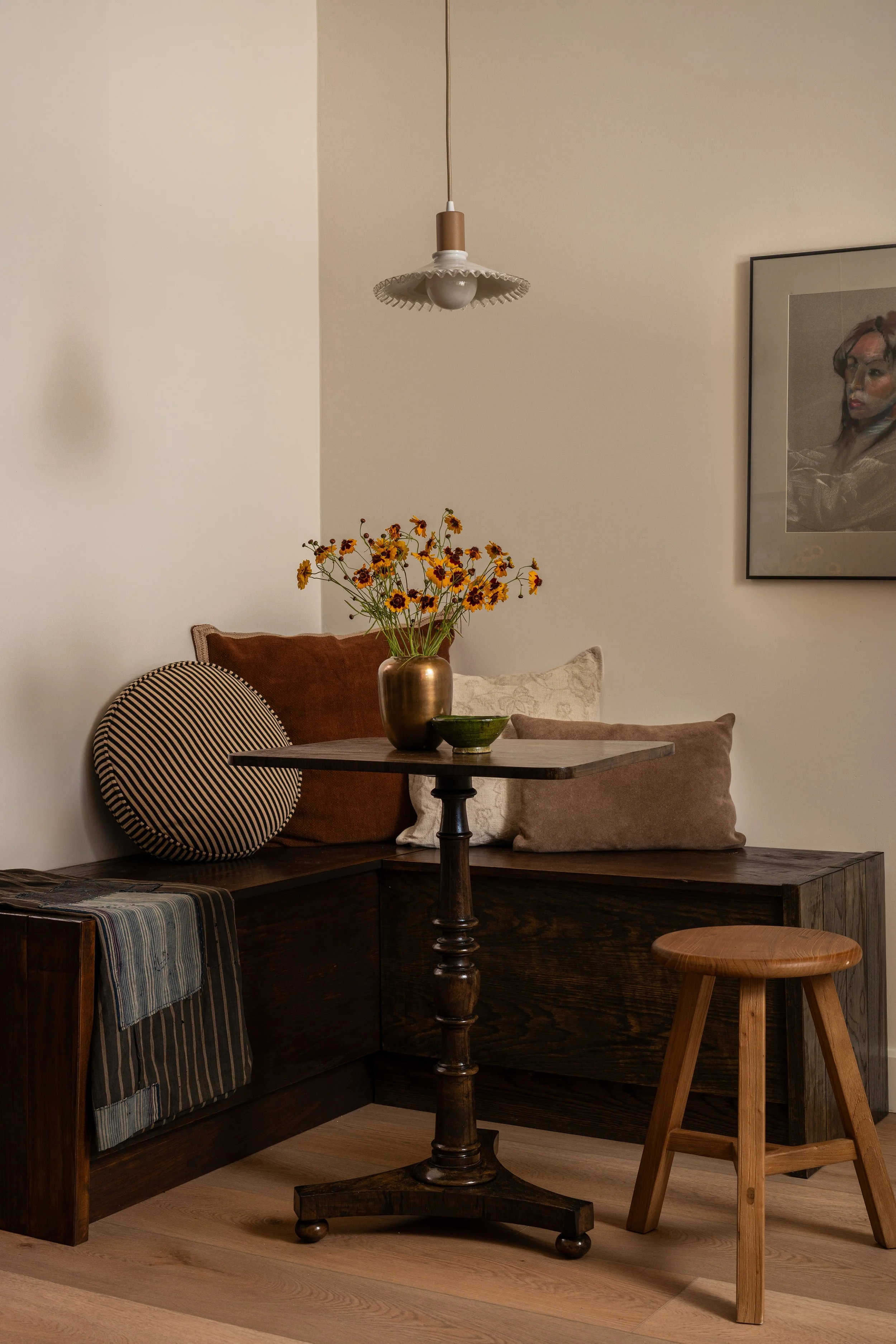 A living room corner with a dark wooden corner bench, decorated with various pillows and a striped blanket. In front, a round wooden table with a dark finish holds a copper vase with yellow and brown flowers and a small green bowl. To the right, a wo