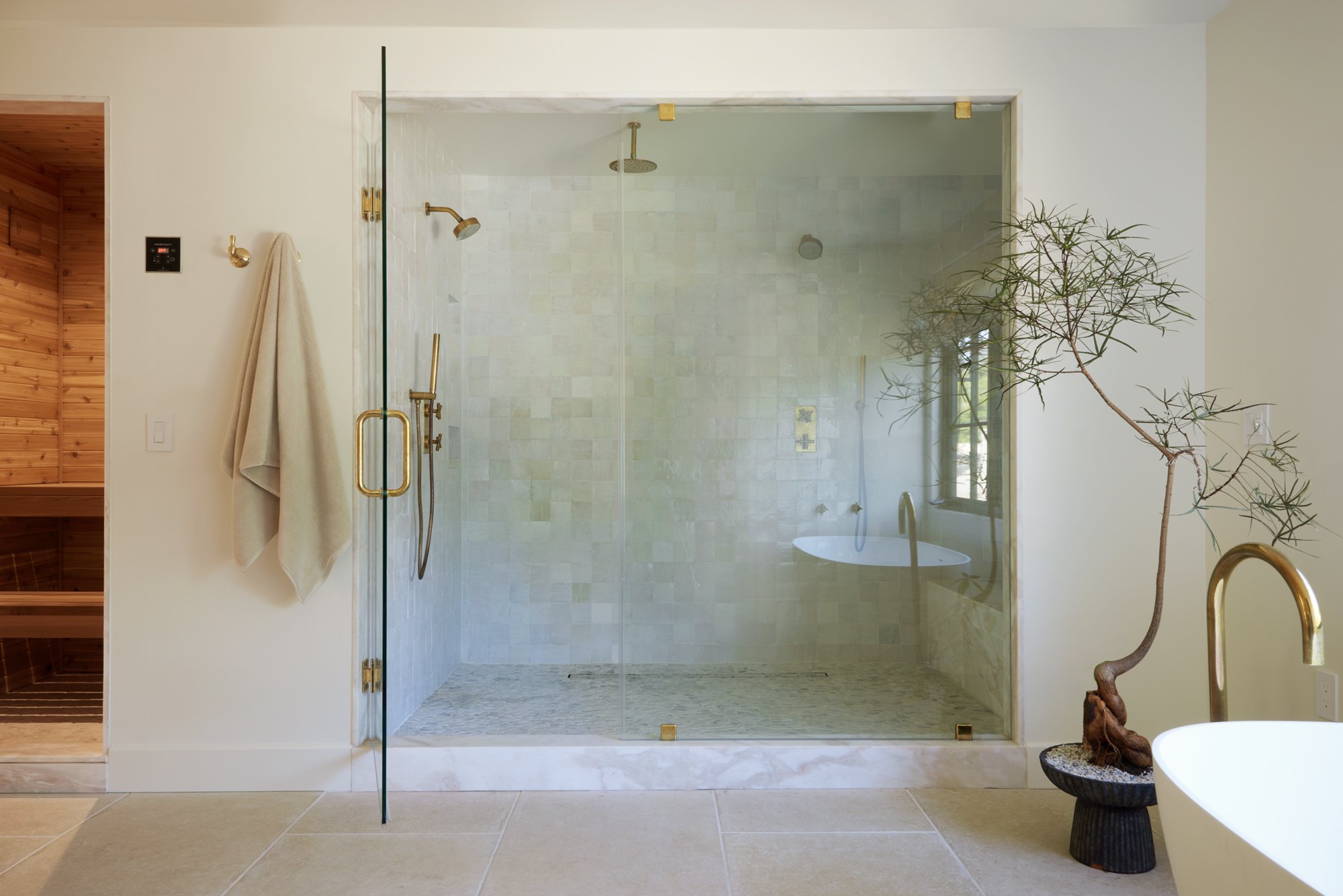A modern bathroom with a walk-in glass shower featuring gold fixtures and beige tiled interior. There is a white wall with two gold hooks and a towel hanging on one. To the right, there is a tall, thin tree in a black pot. To the left, a wooden sauna