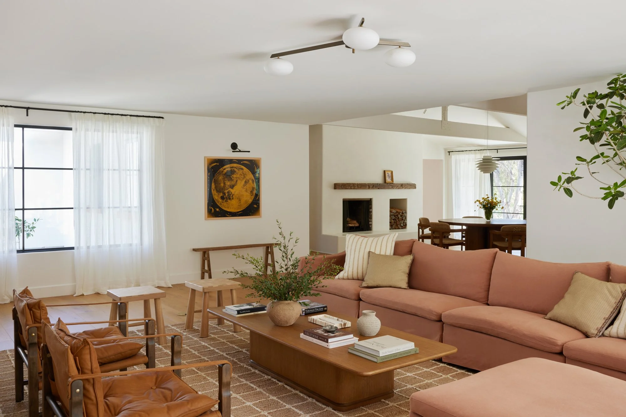 Living room with pink sofa, wooden coffee table with books, armchair with leather cushions, large windows with sheer curtains, artwork of a moon on the wall, and a cozy fireplace.