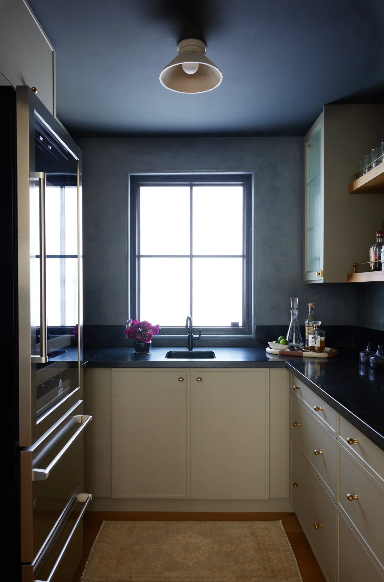 A cozy kitchen with a large window, black countertops, cream cabinets with gold knobs, a vase with pink flowers, and various bottles on the counter. There is a ceiling light and a beige rug on the wooden floor.