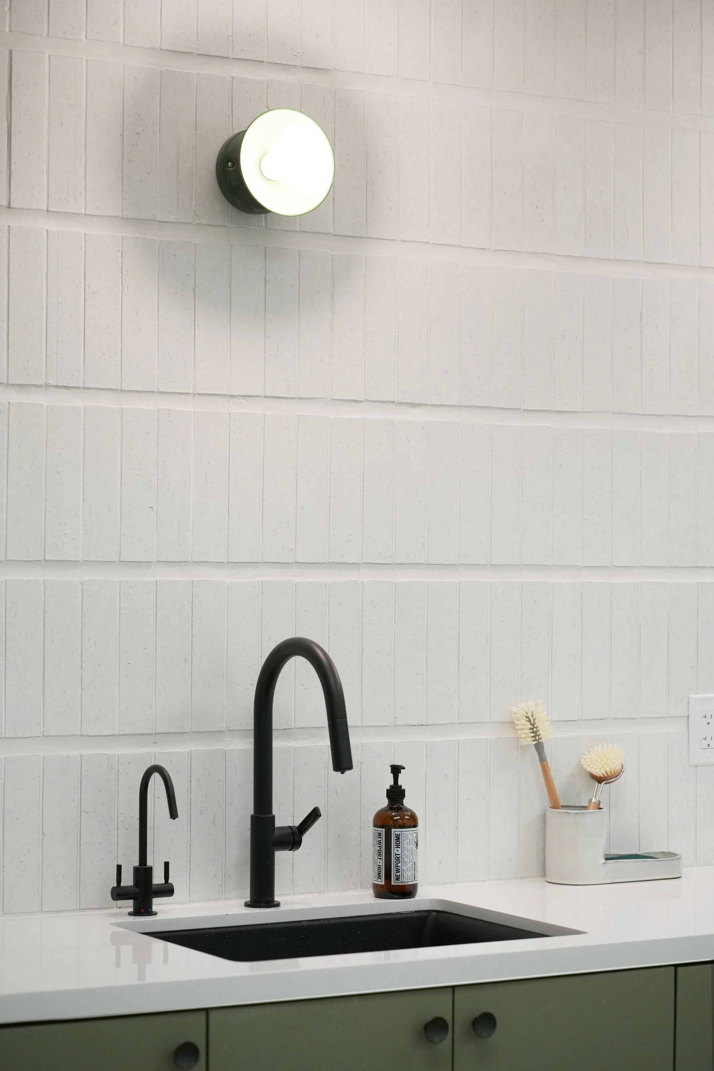 A modern kitchen sink area with black faucet and soap dispenser, and a white countertop. Behind the sink are two brushes in a white container, a brown bottle, and a white tiled wall. There is a light fixture on the wall above the sink.