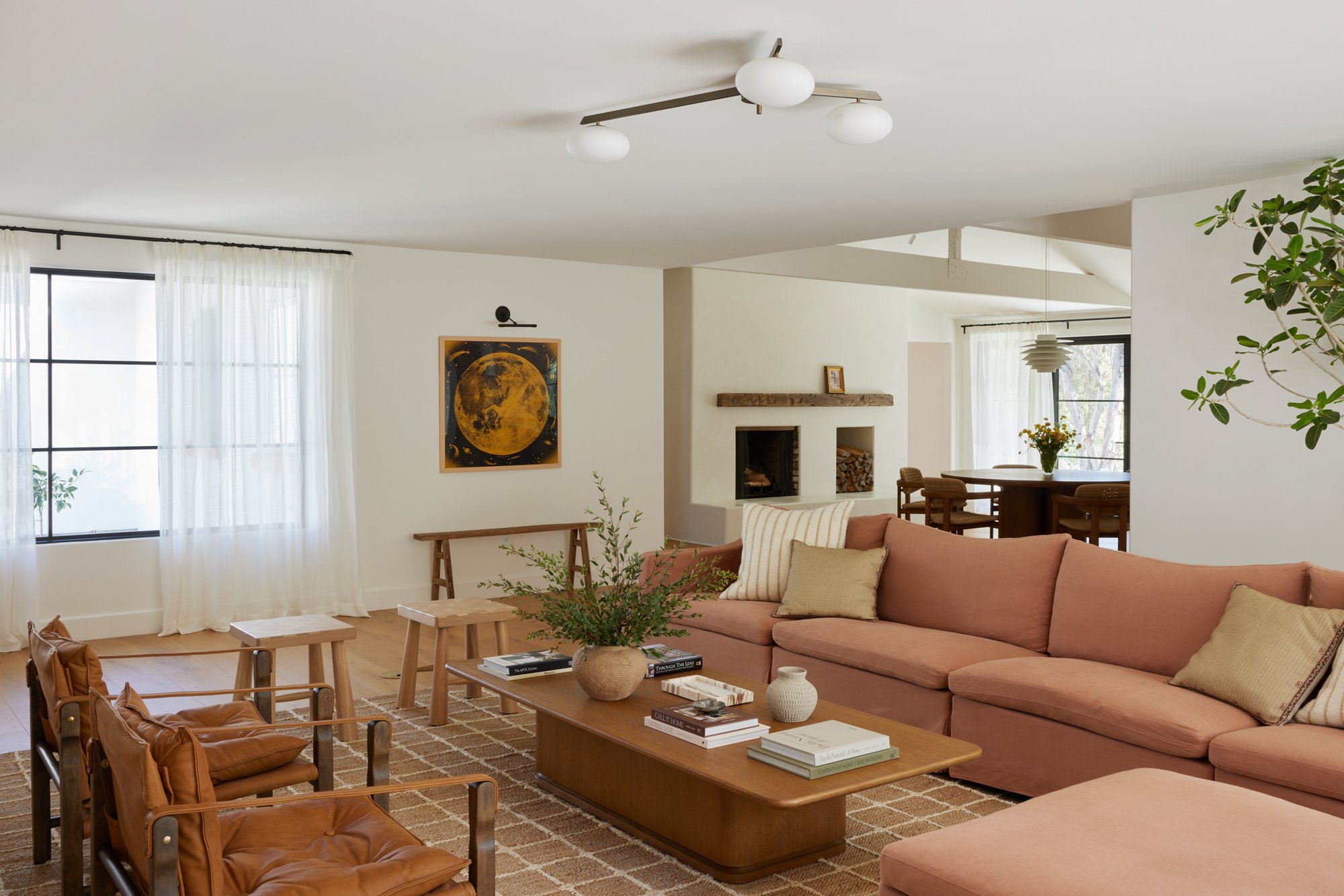 Living room with pink sofa, wooden tables, and armchairs, white curtains, wall art, fireplace, green plants, and windows letting in natural light.
