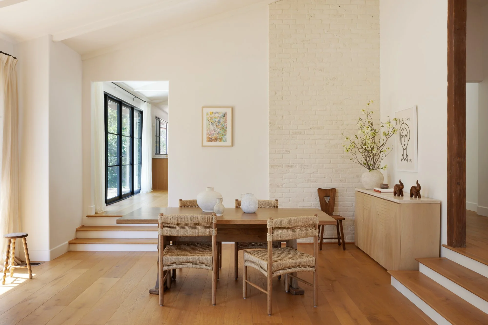 A dining room with a wooden table and six matching chairs. The space features a white brick accent wall, minimalistic artwork, a vase with branches, and decor items. Natural light streams in from large black-framed windows with white curtains, illumi