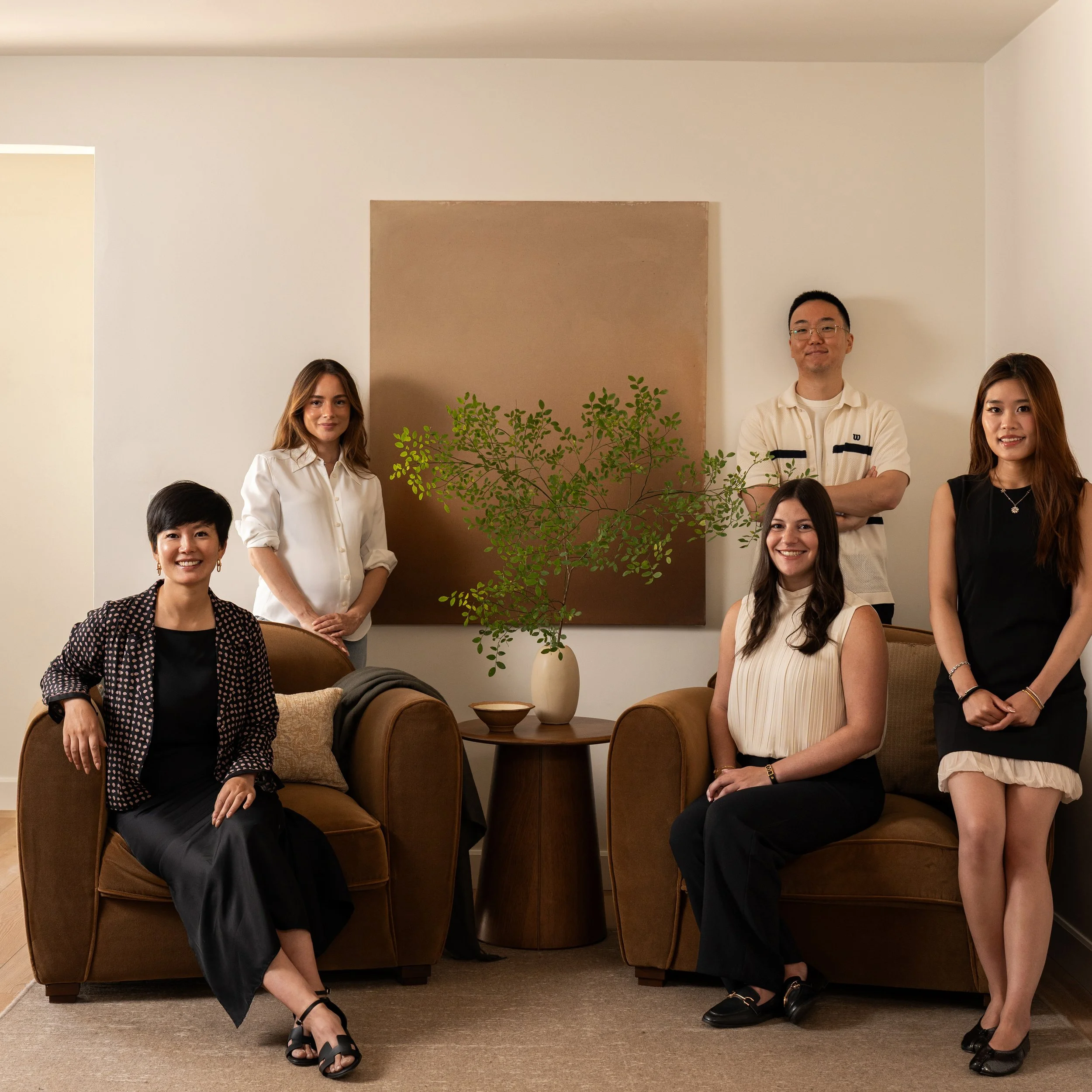 A group of diverse people posing for a photo in a room, with some sitting and some standing, with a plant and artwork on the wall behind them.