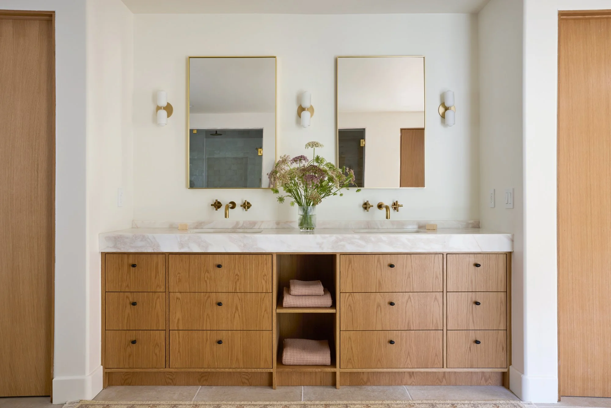 Bathroom vanity with two mirrors, two wall-mounted lights, a vase with flowers, and wooden cabinets.