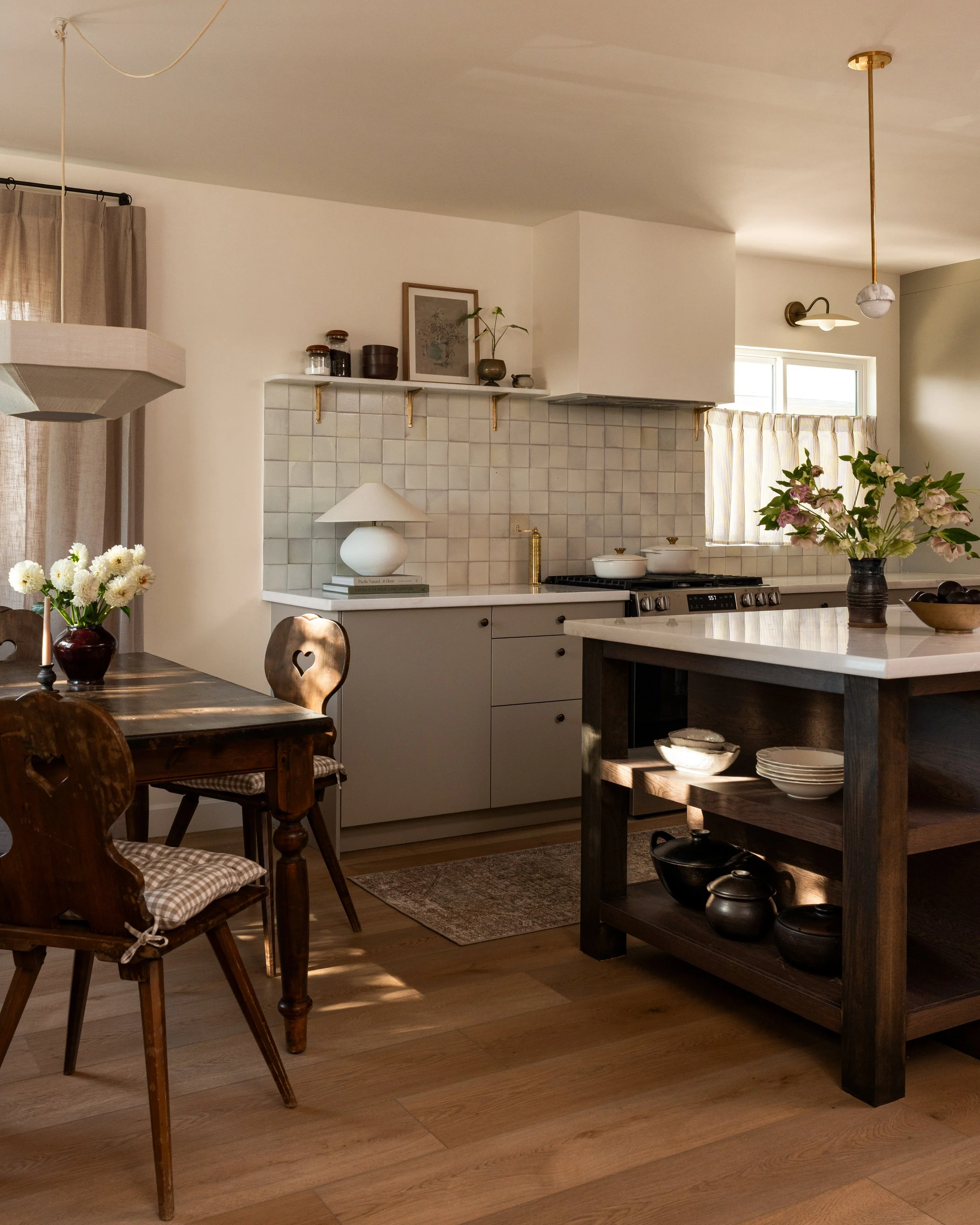 A cozy kitchen and dining area with wooden flooring, a rectangular dining table with wooden chairs, a kitchen counter with white cabinets, a black wooden island with bowls, and decorative items. Flowers in a dark vase are on the table, and shelves with jars and artwork are on the wall. Natural light comes through windows, and ceiling pendant lights add warm lighting.
