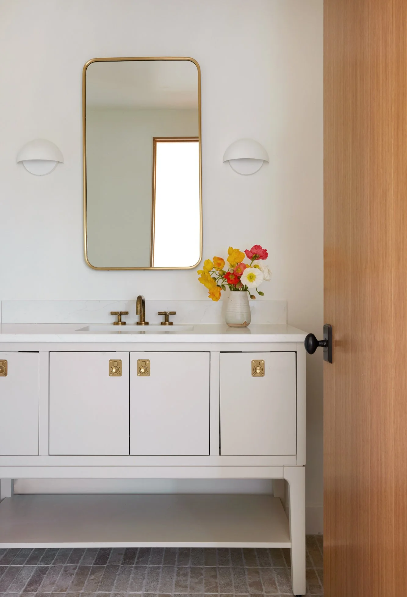 A bathroom vanity with white cabinetry, gold hardware, and a white marble countertop with a gold faucet. Above it, a tall rectangular mirror with a gold frame, flanked by two white wall sconces. To the right, a vase of colorful flowers sits on the co
