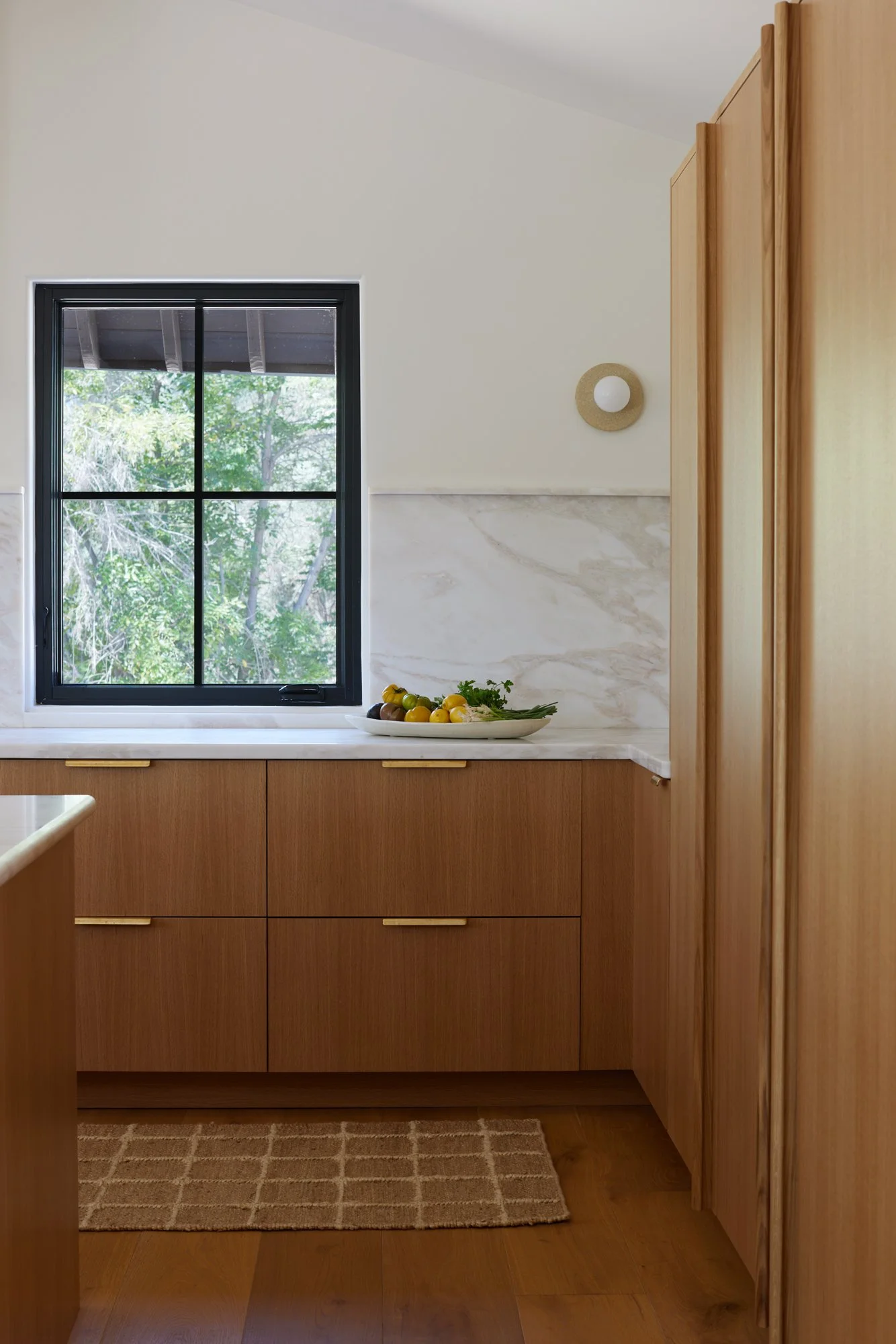 Kitchen with a large window showing green trees outside, wooden cabinets, a marble countertop, a decorative wall light, a beige rug, and a dish with tomatoes, lemons, and herbs.