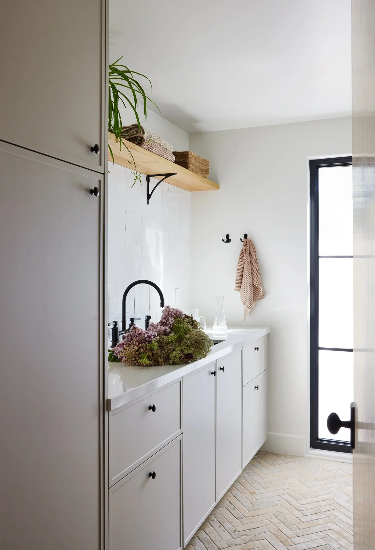A bright white laundry room with a white countertop, black hardware, and a black-framed glass door. There is a wooden shelf with baskets and towels above the sink area. A black faucet, a vase, and a bouquet of flowers are on the countertop. Two black