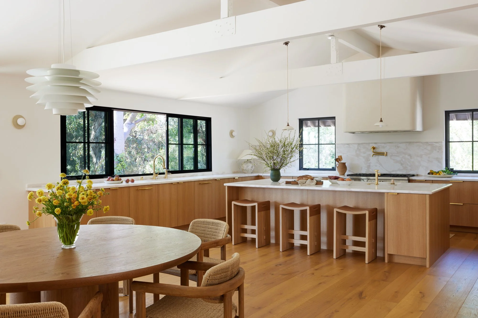 A modern kitchen with wooden cabinets, a marble island, black-framed windows, pendant lights, and a dining area with a round table and woven chairs.