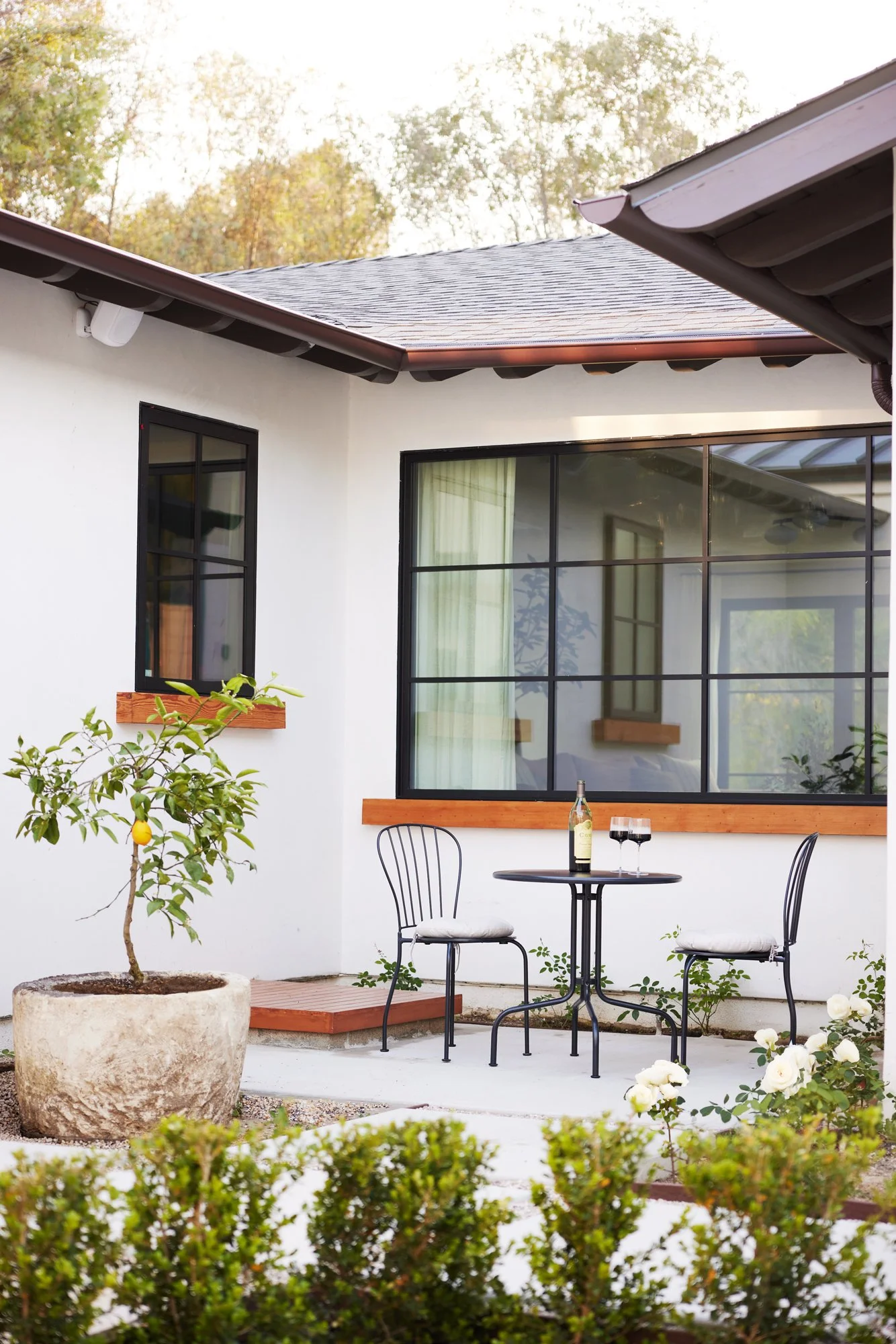 A cozy outdoor patio scene with a black metal table and two matching chairs with white cushions. On the table, there's a bottle of wine and two glasses filled with red wine. To the left, there's a potted lemon tree, and in the background, a white ext