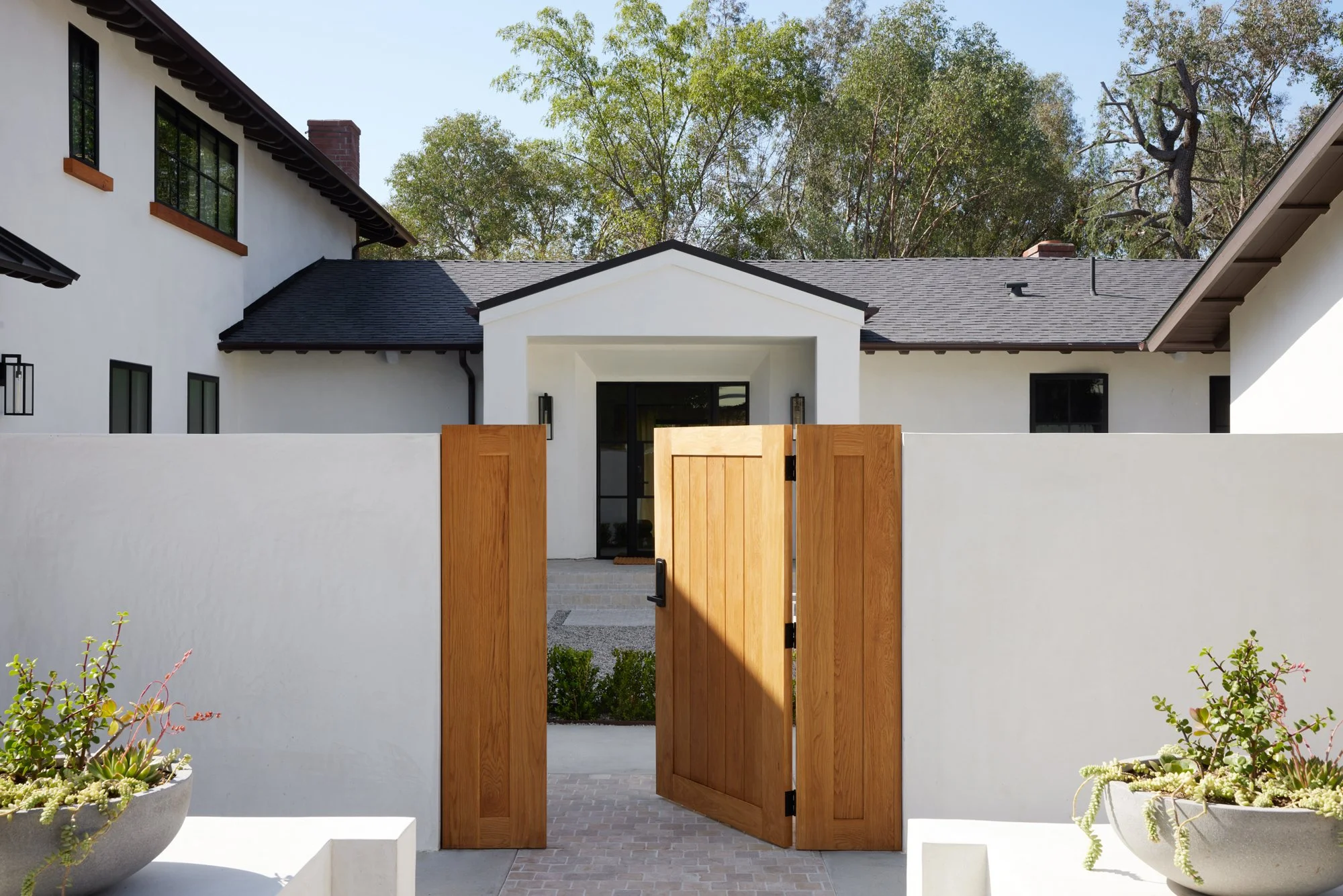 A modern white house with black roof shingles, black window frames, and a wooden front door, set behind a white wall with a wooden gate. There are potted plants with succulents in front of the wall and trees in the background.