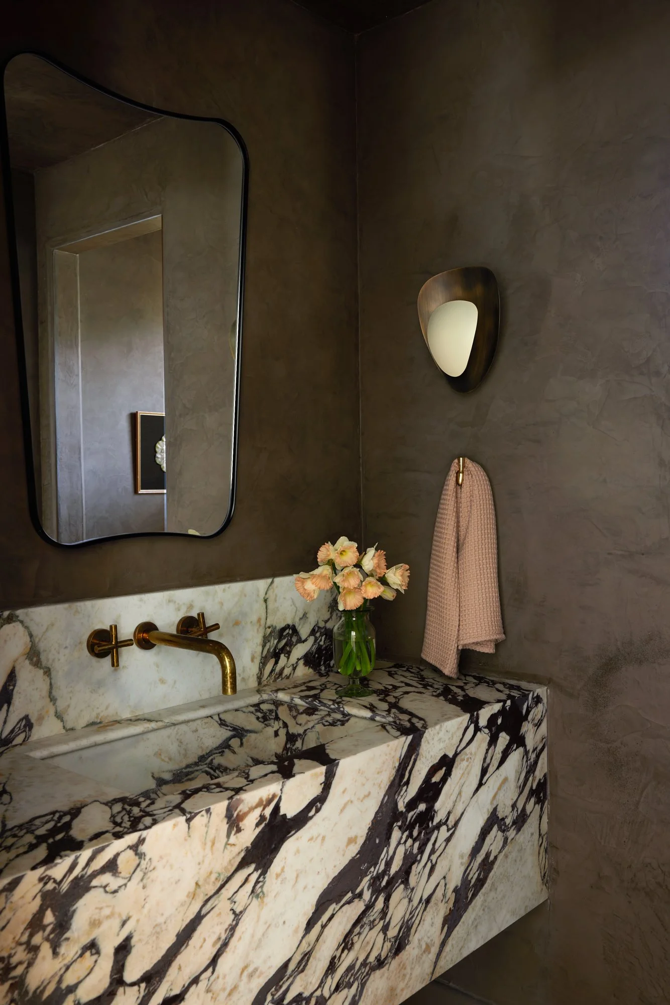 A bathroom vanity with a black and white marble countertop, a large rectangular mirror, a wall-mounted gold faucet, a vase with pink flowers, a round wall light, and a pink towel hanging on the wall.