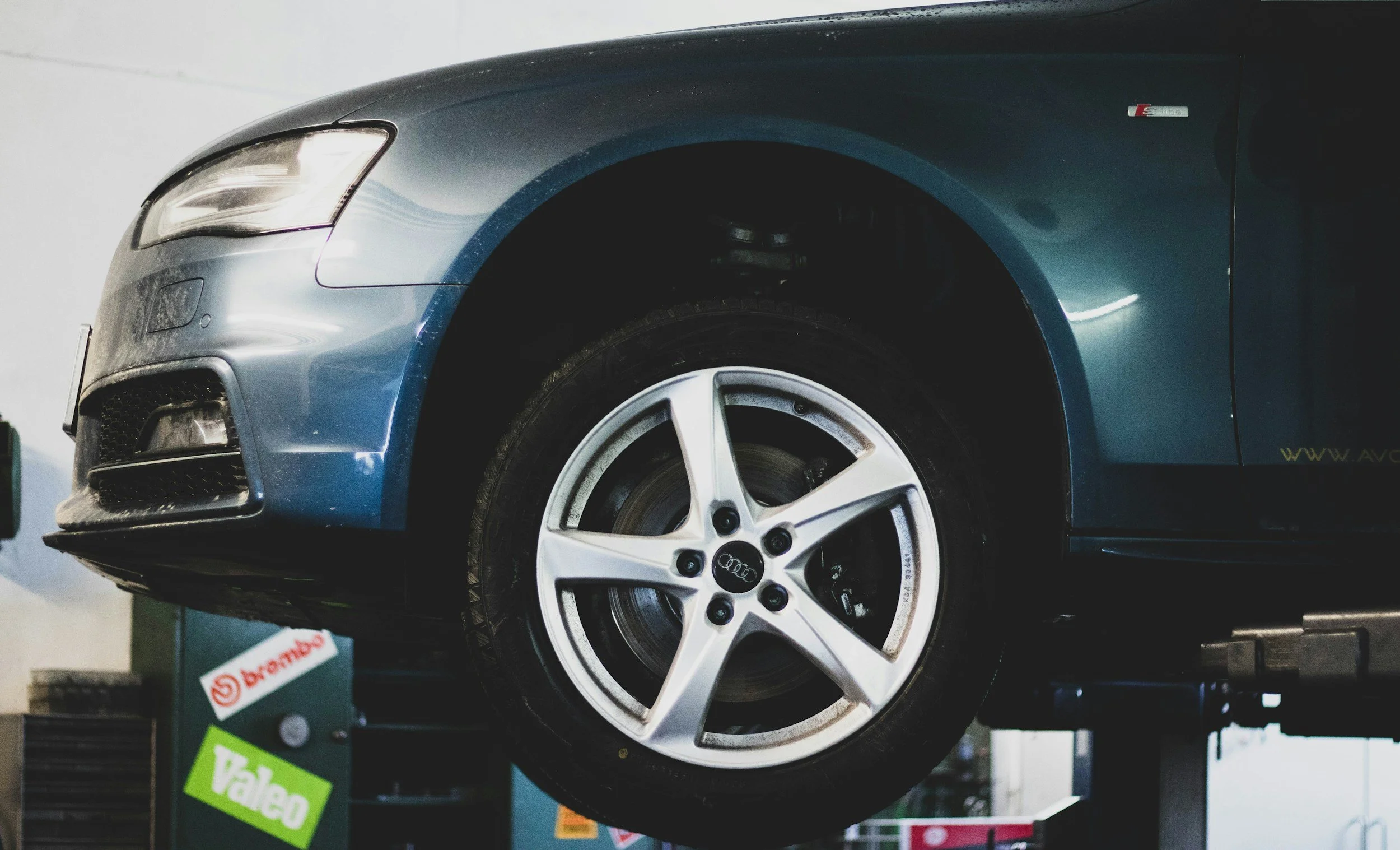 Close-up of a metallic blue Audi car on a hydraulic lift in an auto repair shop, showing the front wheel and part of the front bumper.