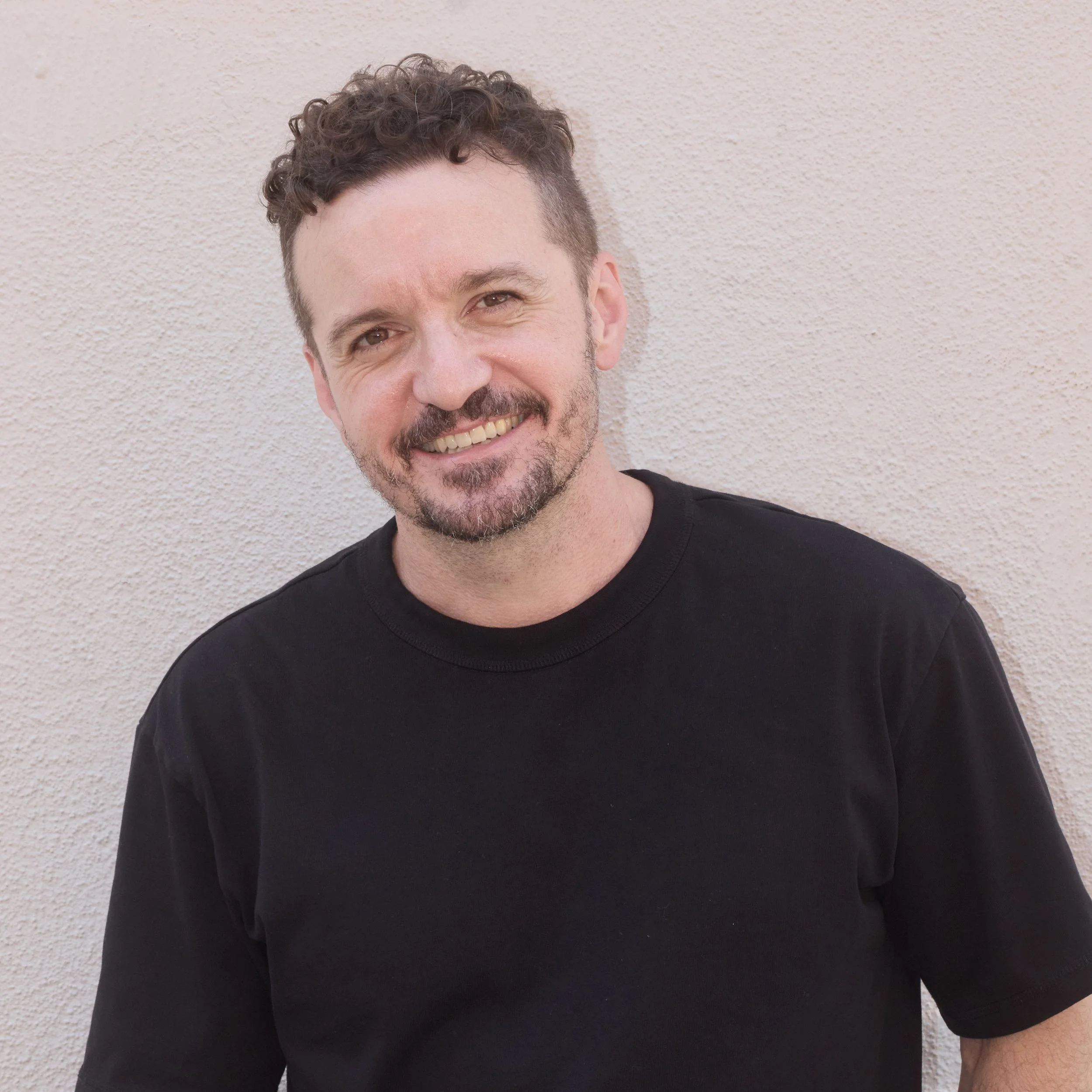 A male professional photographer with short curly brown hair and a beard, smiling, wearing a black t-shirt, standing against a textured beige wall.