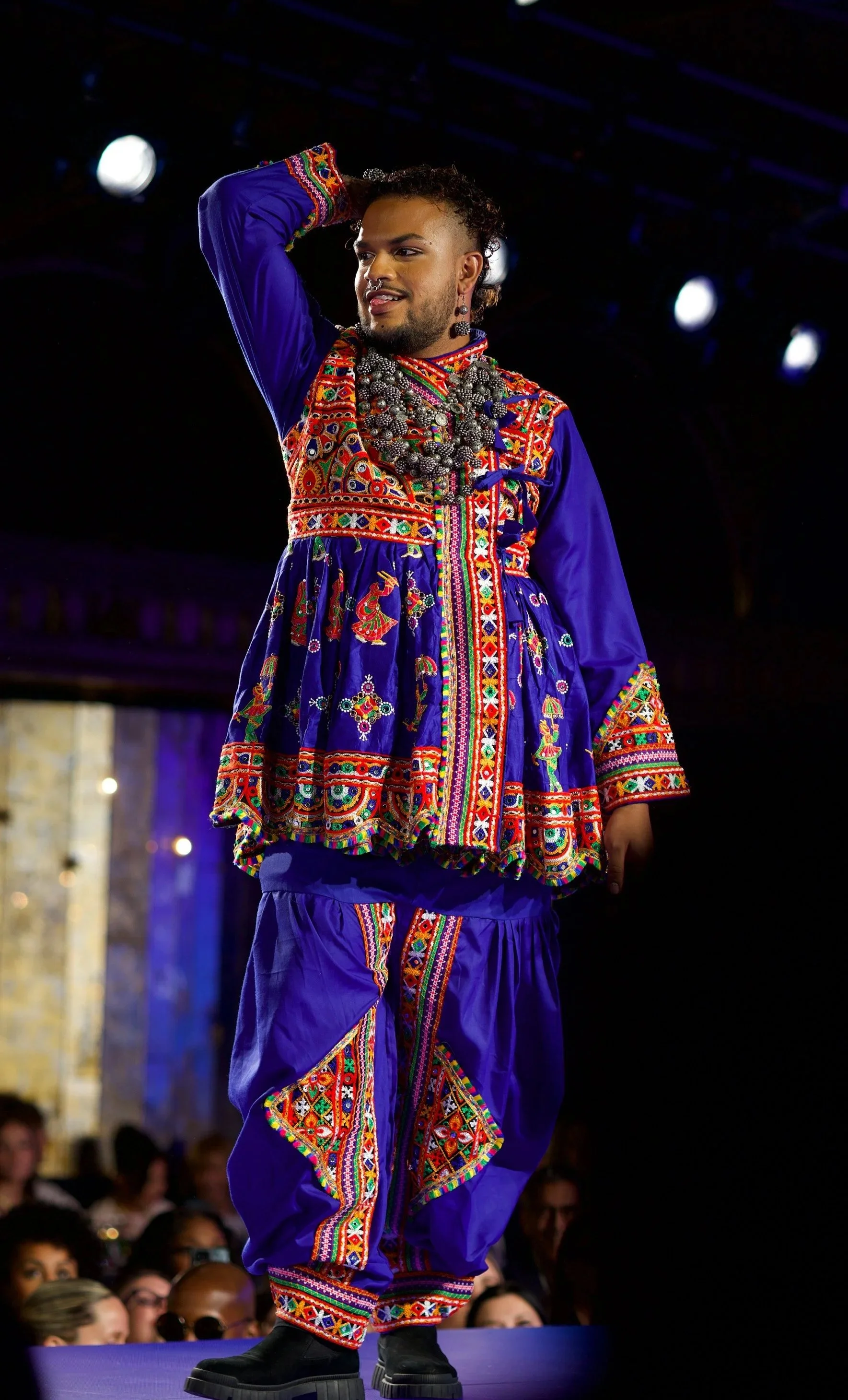 A man fashion model, modeling traditional elaborately embroidered clothing with a matching necklace, standing on a runway at a fashion show in front of an audience.