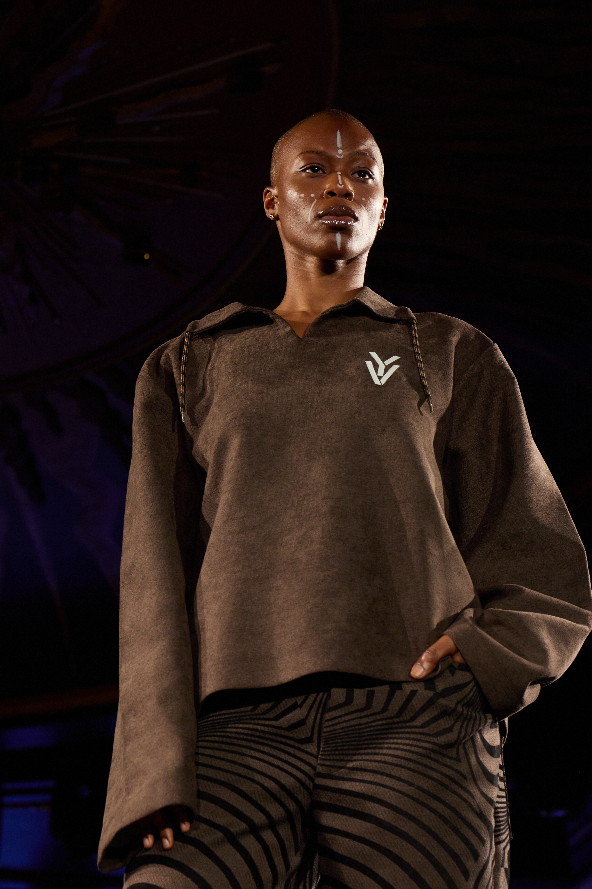 A woman fashion model with a shaved head wearing a brown sweatshirt with a white logo and striped pants, standing confidently on a runway with a dark background.