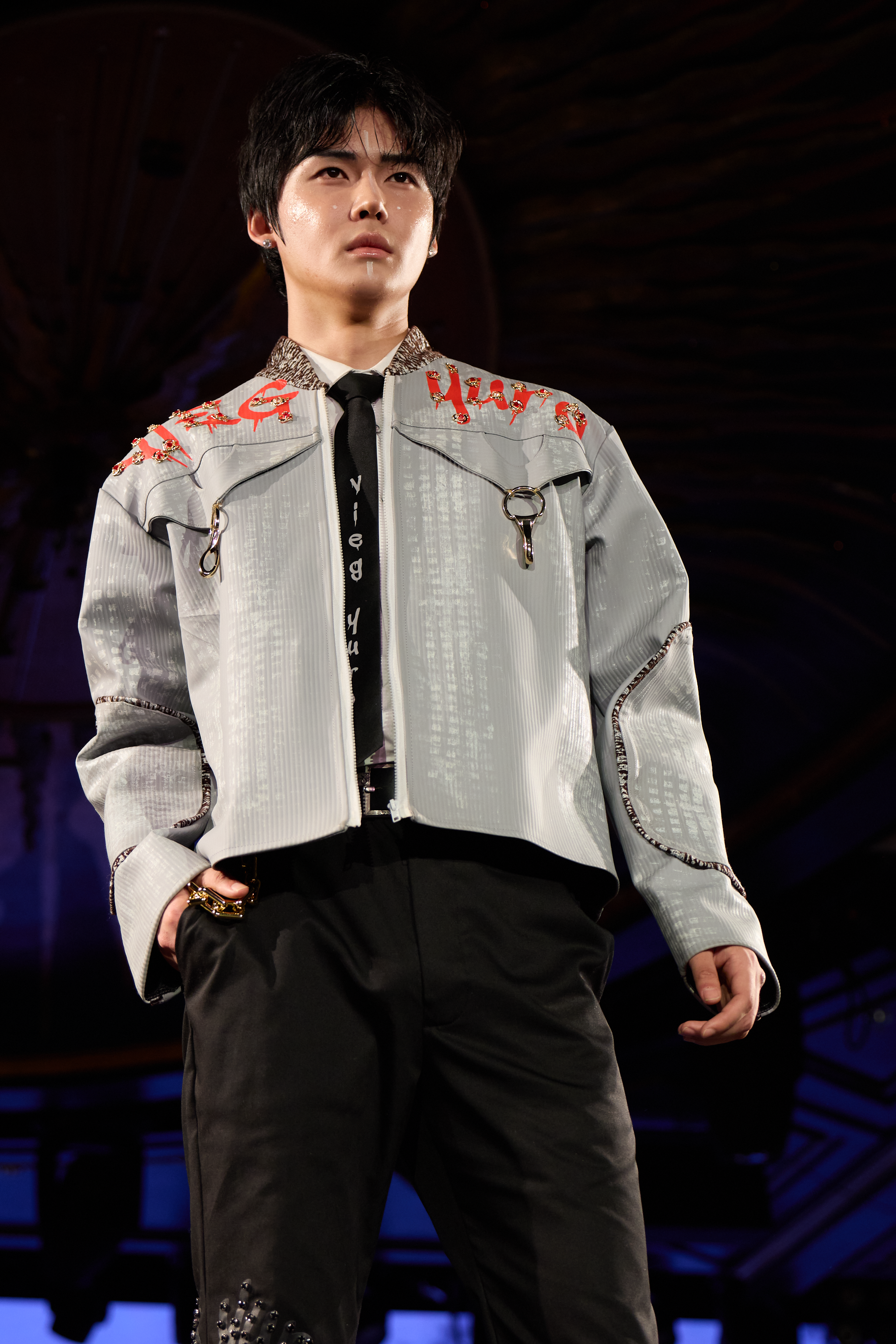 A male model on a runway during a fashion show, wearing a gray jacket with decorative red embroidery on the shoulders, a black shirt with white text, and black pants with studded details.