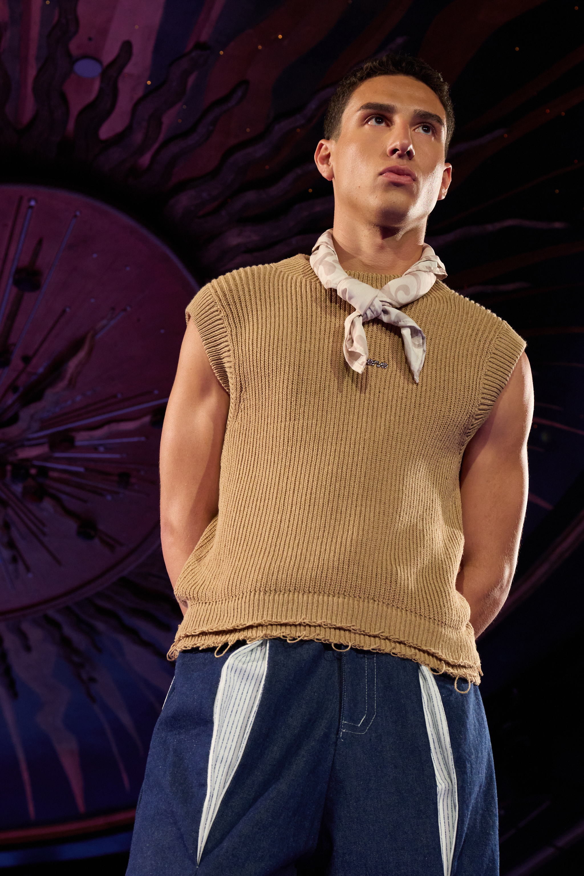 A young male fashion model with short dark hair and a serious expression, wearing a sleeveless beige knit top, a white patterned scarf, and loose-fitting dark blue jeans standing on a runway with dark, artistic background with purple and pink hues.