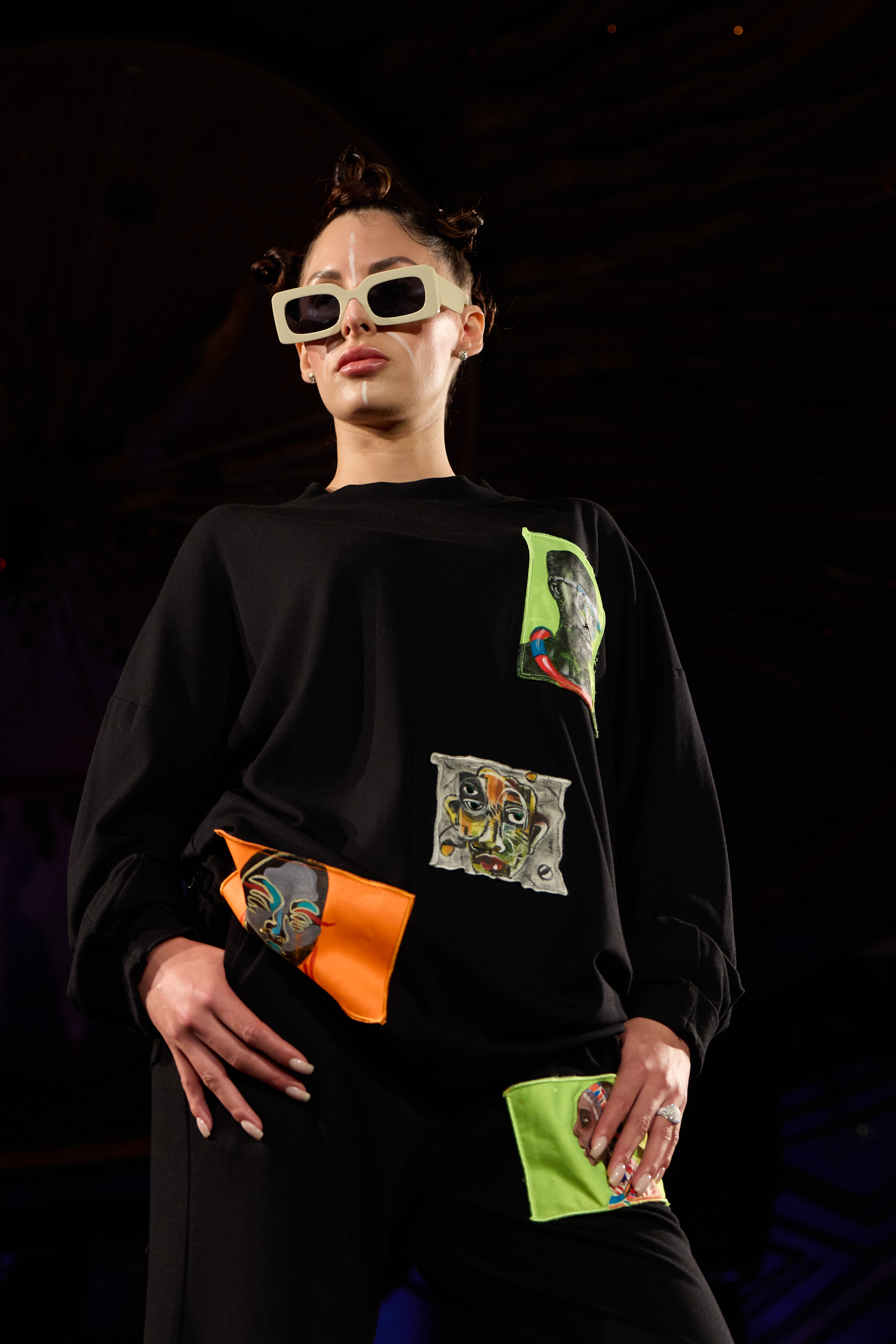 Female fashion model wearing large sunglasses, black top with colorful abstract patches, and matching accessories on her clothes, on a runway with dark lighting.
