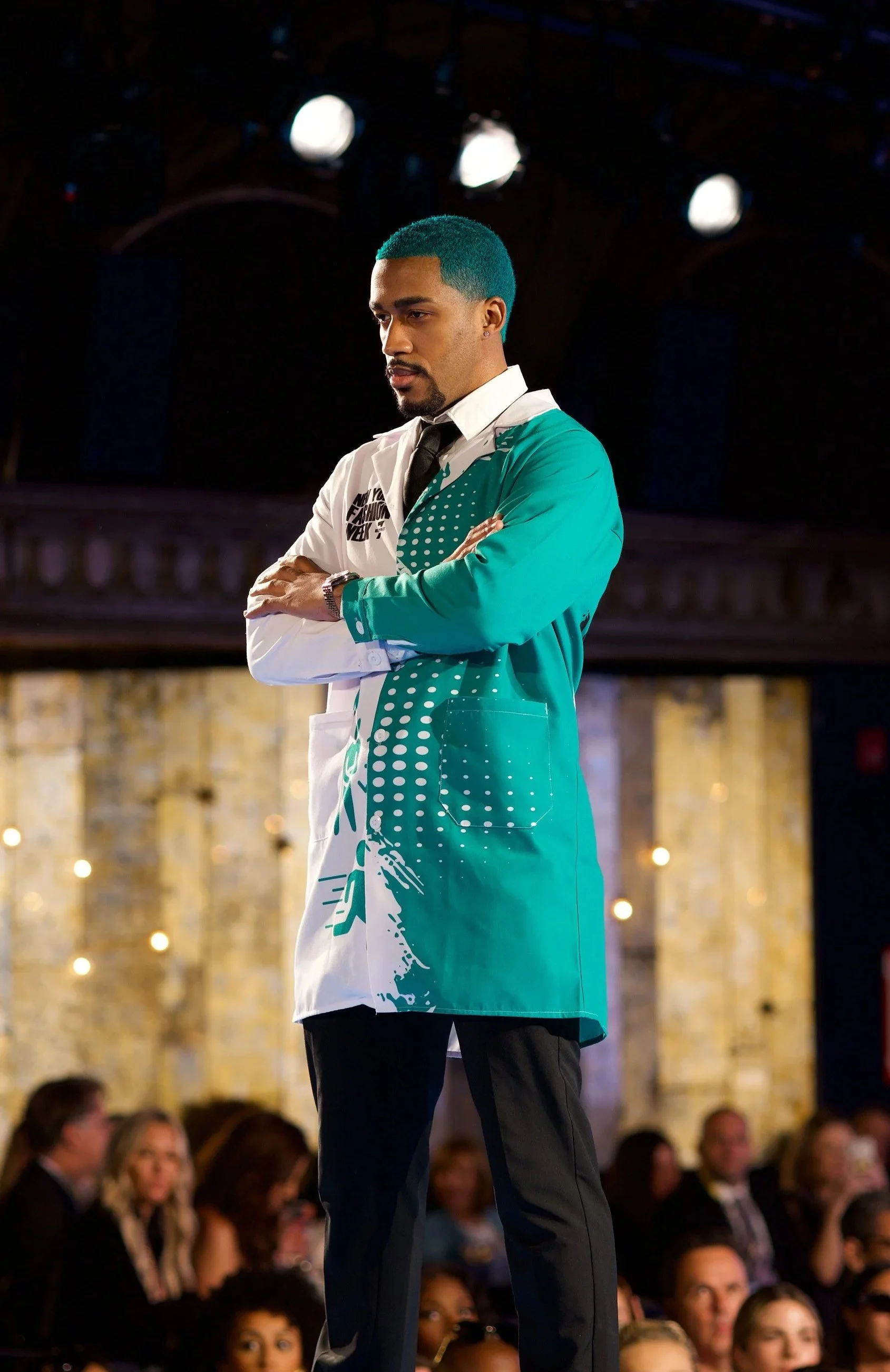 A male fashion model with turquoise hair and a colorful fashionable custom lab coat stands with arms crossed on a runway stage at a fashion event, with an audience in the background.