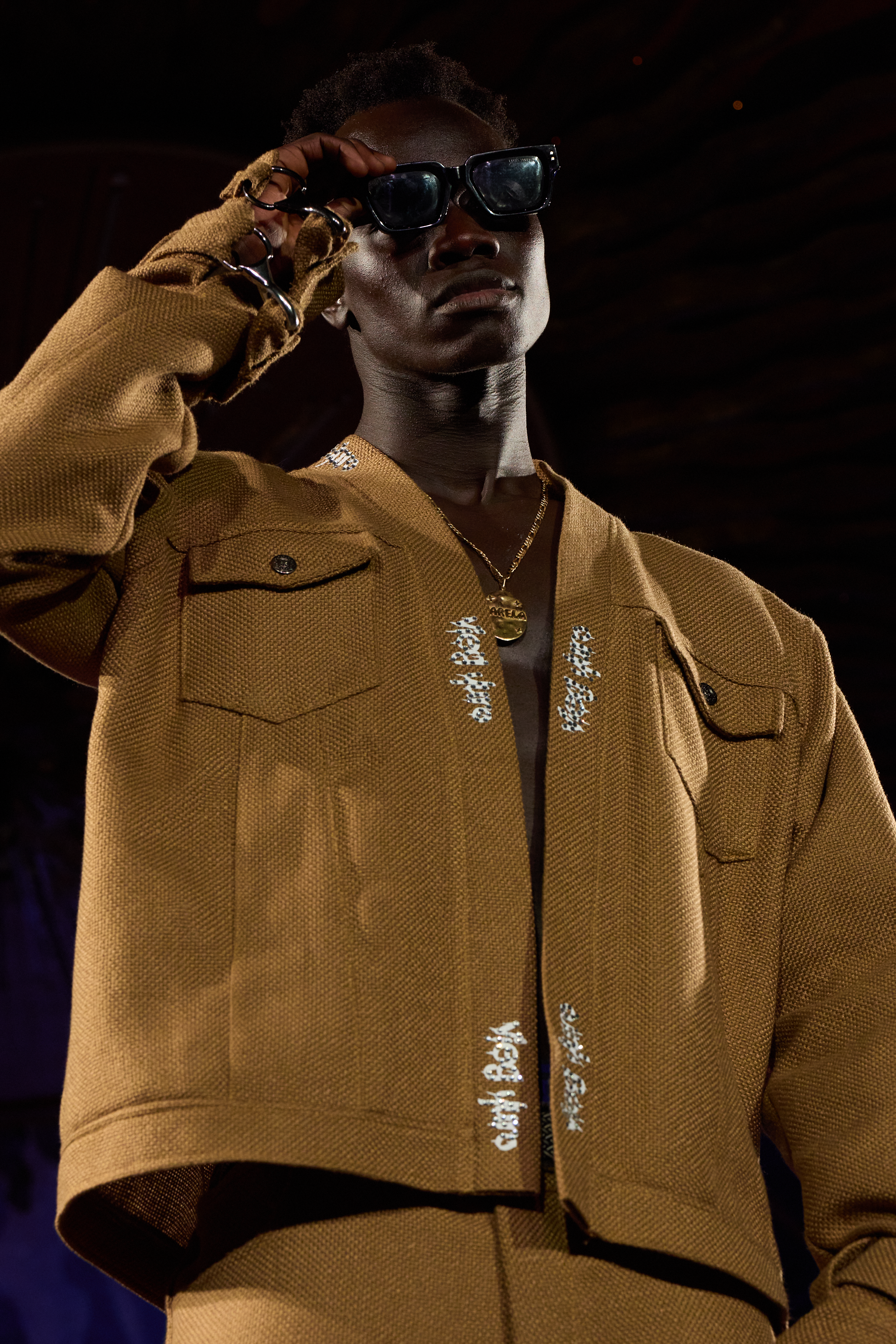A male model wearing a brown jacket, black sunglasses, and a gold chain with a pendant, on a runway at a fashion show and looking confidently at the camera.