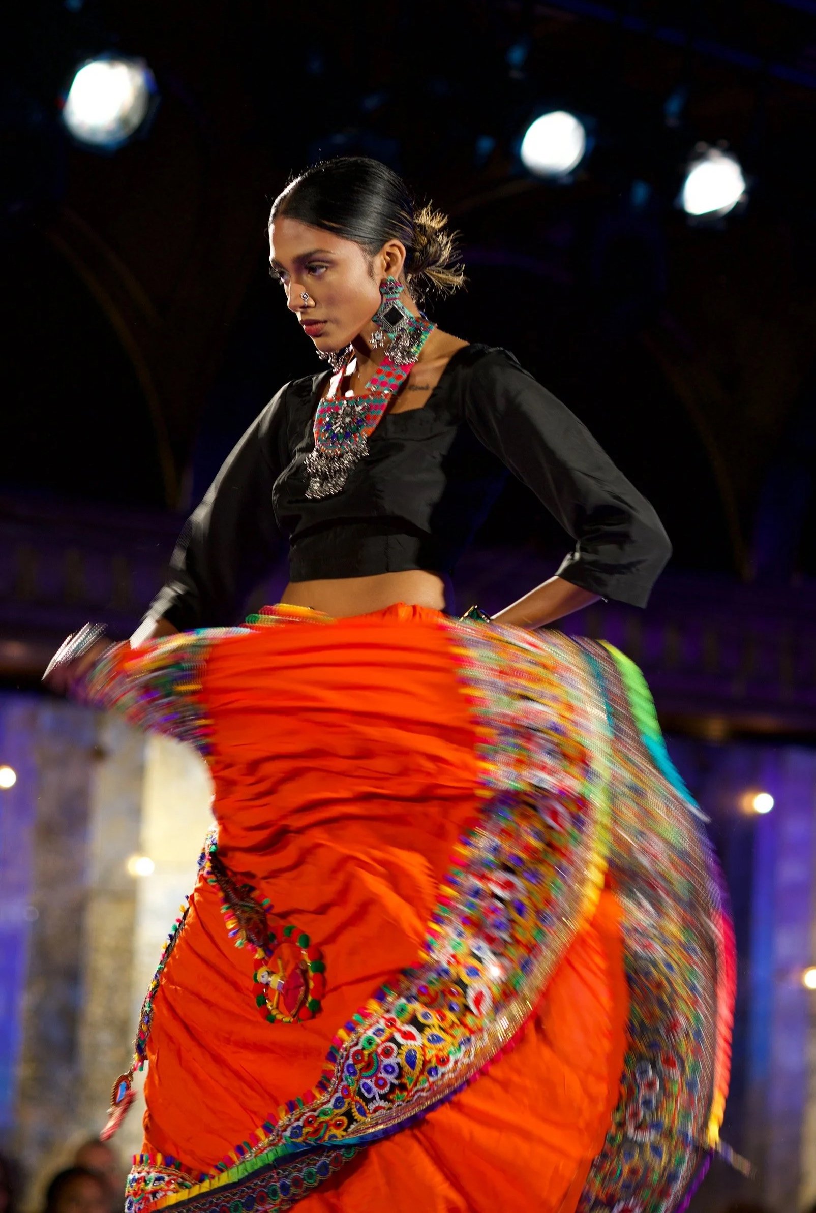A woman wearing a black top with 3/4 sleeves, colorful jewelry, and a vibrant orange skirt with embroidered patterns, dancing on a dimly lit runway at a fashion show with stage lights in the background.
