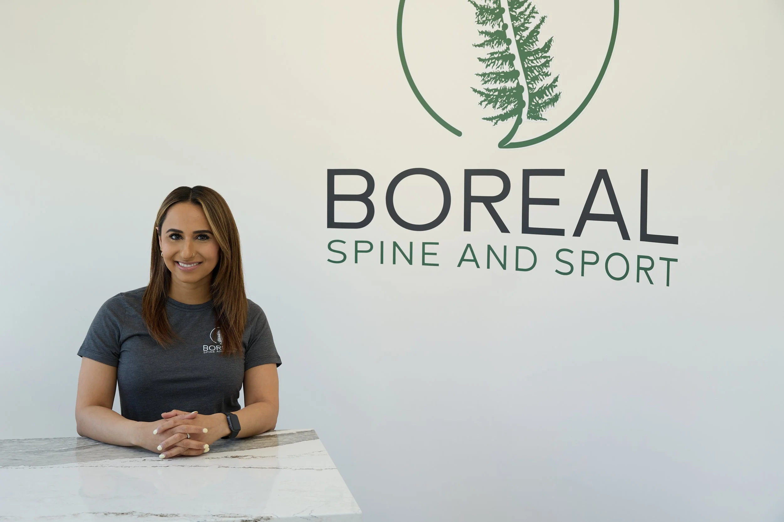 A woman standing with arms crossed, smiling, in front of a sign that reads 'Boreal Spine and Sports' with a tree logo.
