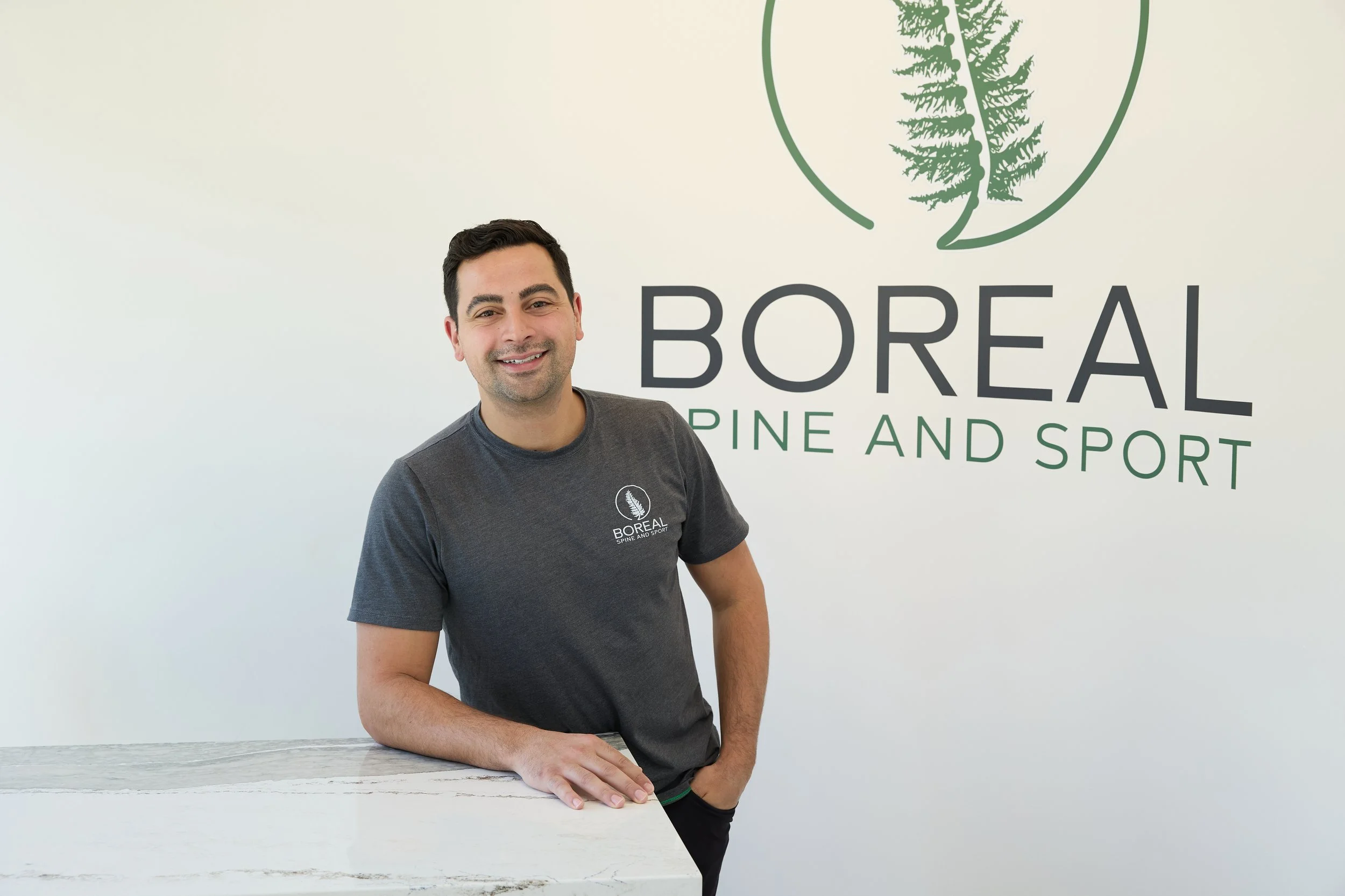 A smiling man in black scrubs sitting on a therapy table in a medical office with white walls, green accent wall, and medical charts.