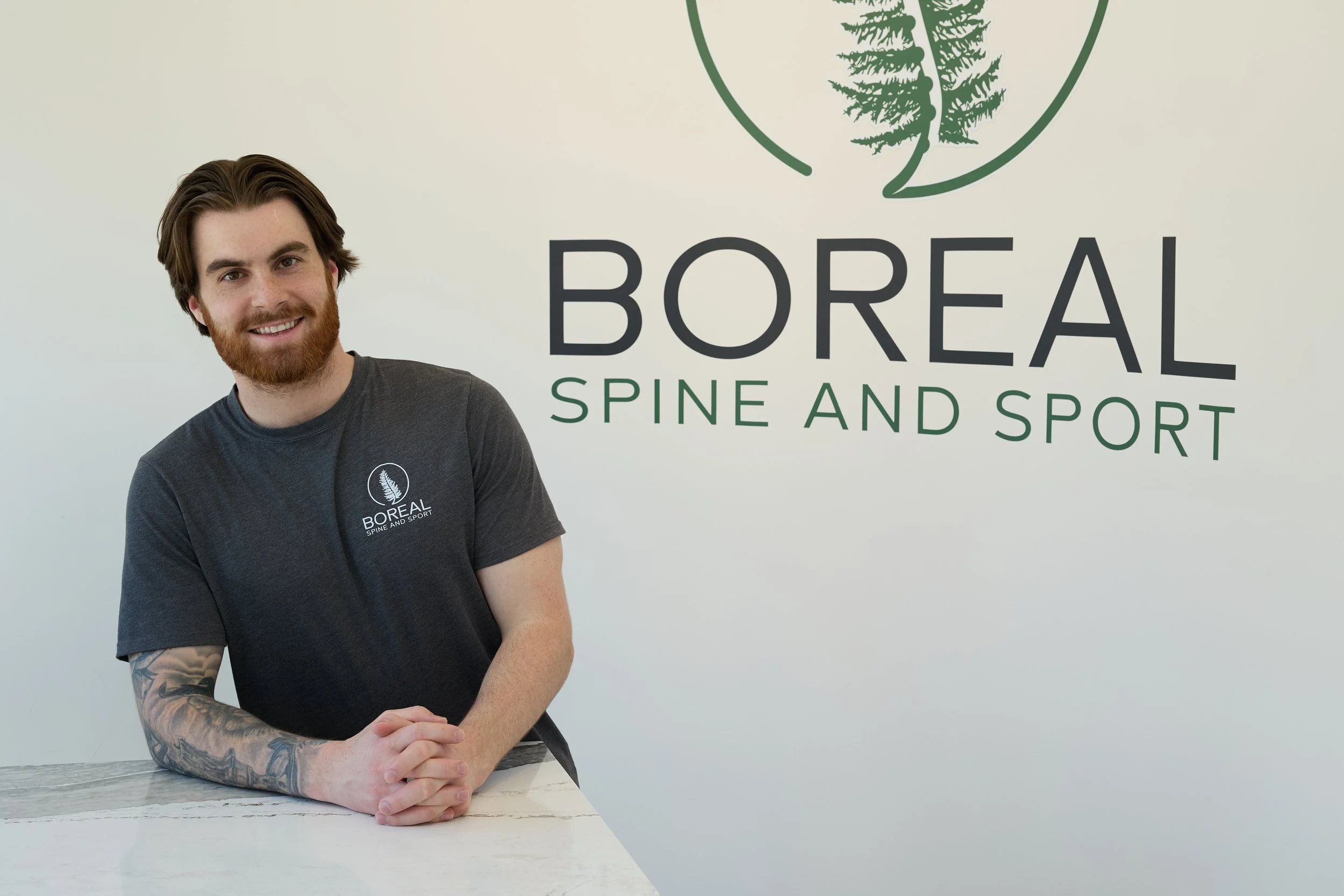 A smiling man with a beard, wearing a black polo shirt and blue pants, standing in front of a wall with the logo of 'Boreal' and a tree graphic, in an indoor setting.