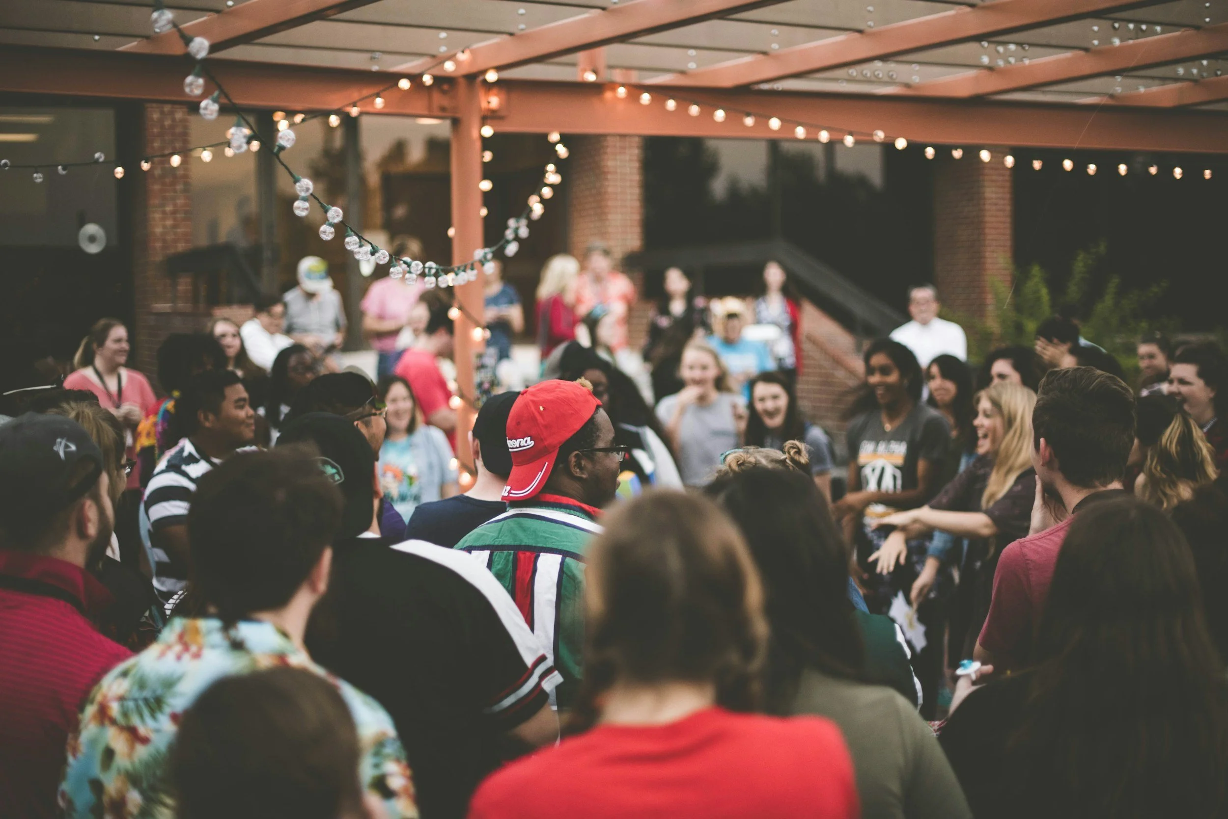 A large gathering of people outdoors under a wooden pergola with string lights at dusk or evening, some people smiling and engaged in conversation.