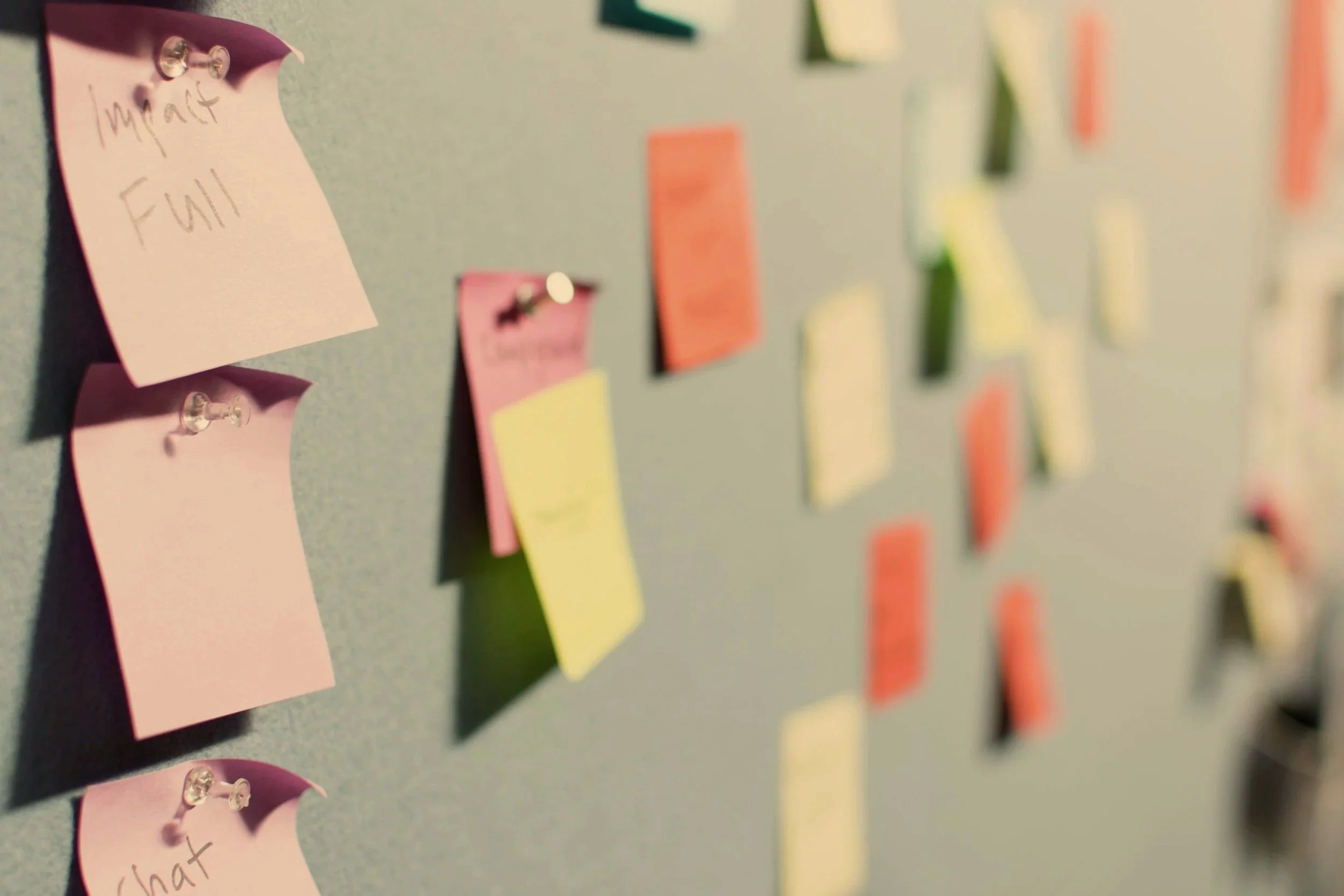 Close-up of a bulletin board with pastel-colored sticky notes and push pins. The notes have handwritten text such as 'what full' and 'what.' The focus is on a pink sticky note with the words 'what full' by a small metallic push pin, while the background shows more sticky notes and push pins out of focus.