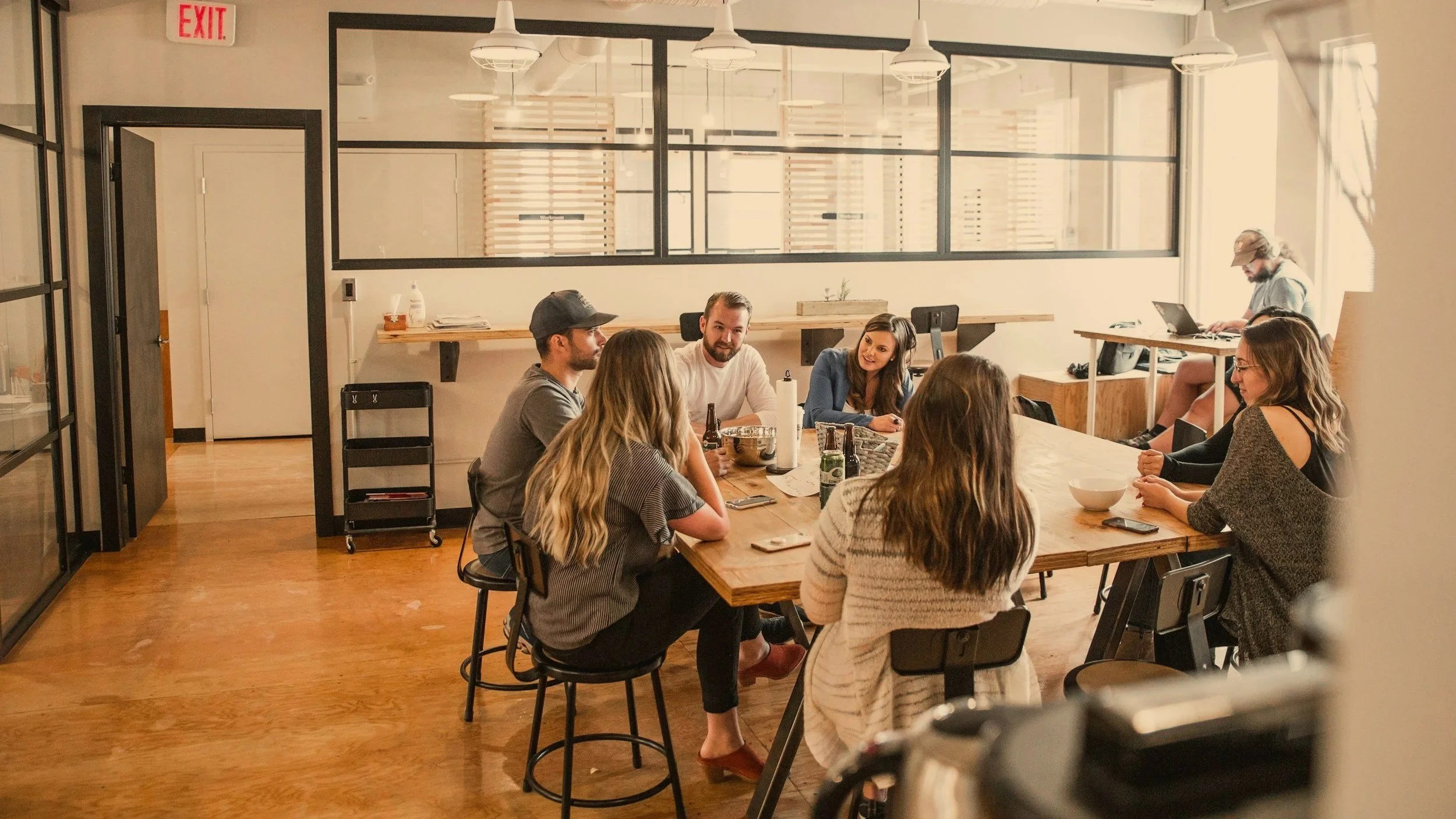 A group of six people sitting around a wooden table in a modern office space, engaged in conversation. In the background, a person is working on a laptop at a separate desk near a window. The office has an industrial design with exposed ductwork, pendant lighting, and glass partitions. There are bottles and a bowl on the table.