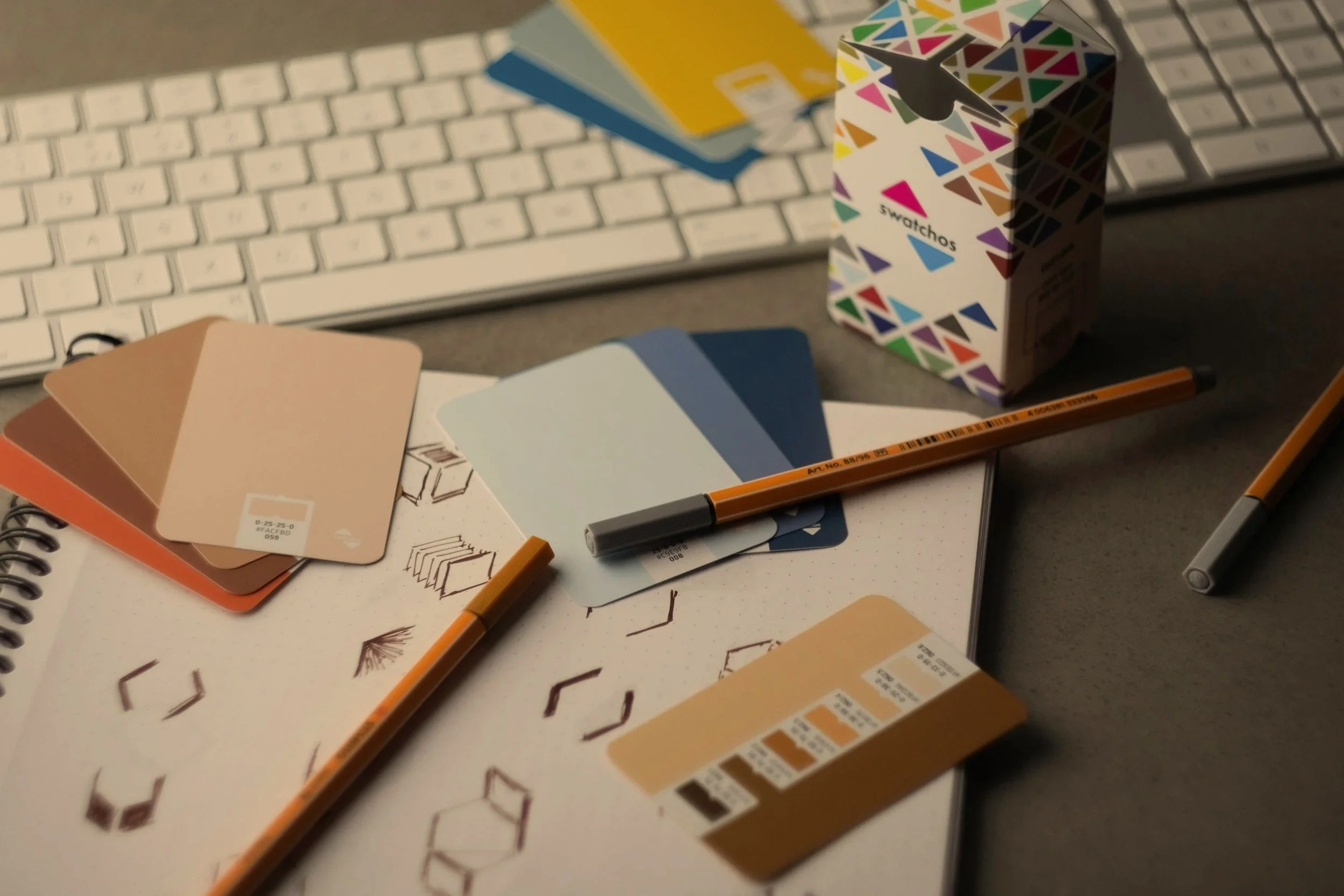 Color swatch sample book, paint color cards, coloring pencils, and decorative box placed on a desk with a keyboard in the background.