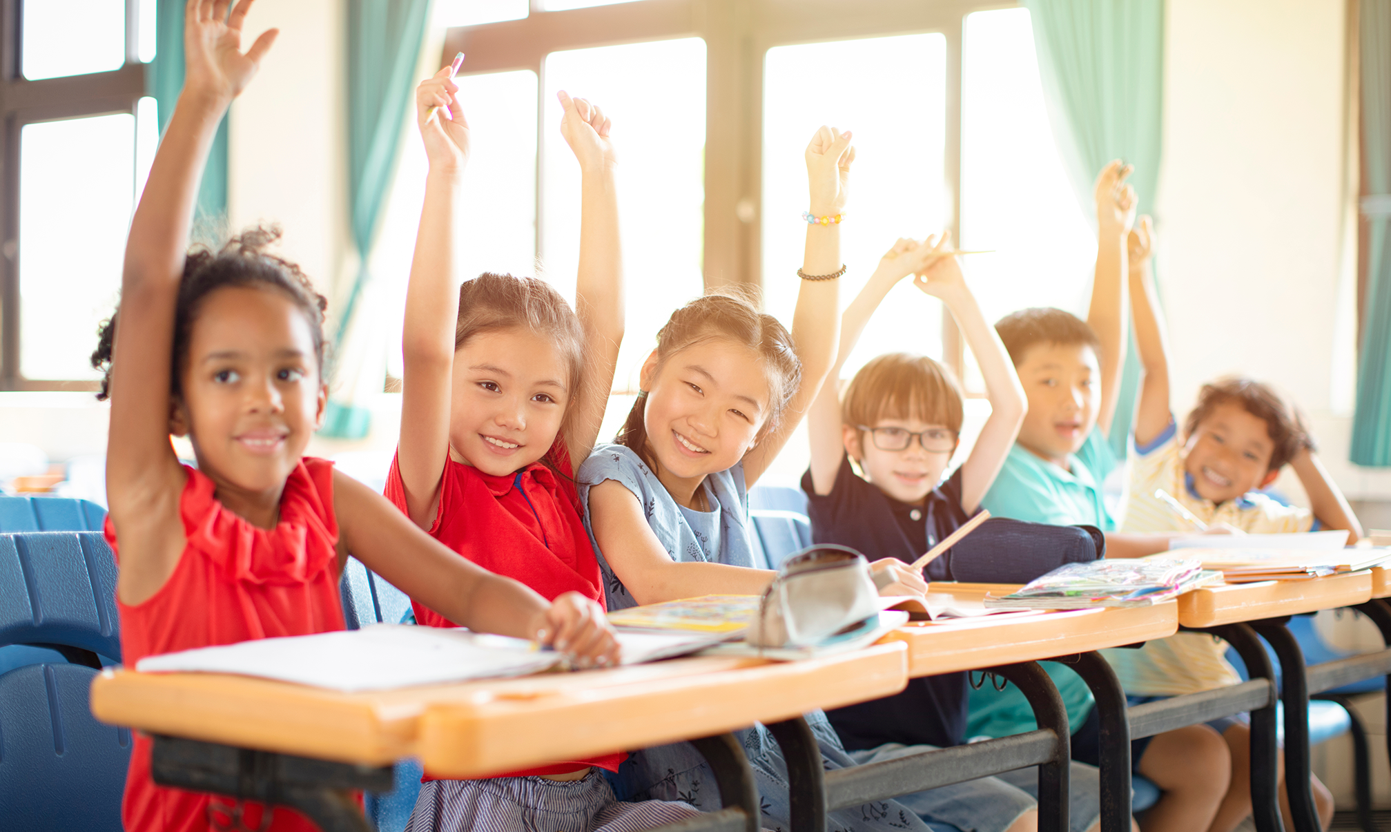 Six young students sit at desks all raising their hands